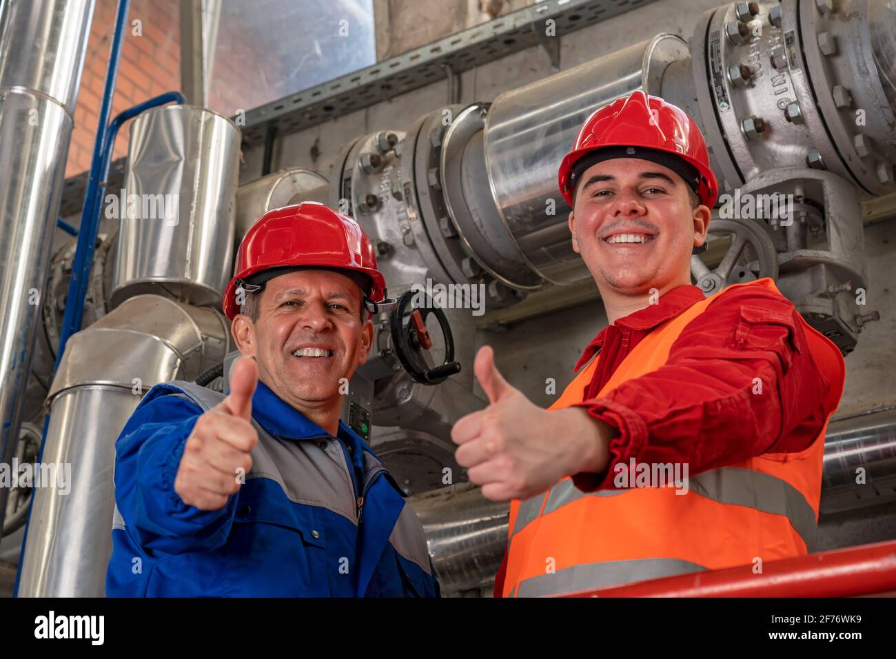 Lächelnder Öl- und Gasingenieur und junger Arbeiter in Red Coveralls und HardHat, der neben der industriellen Rohrleitung Thumbs gibt. Stockfoto