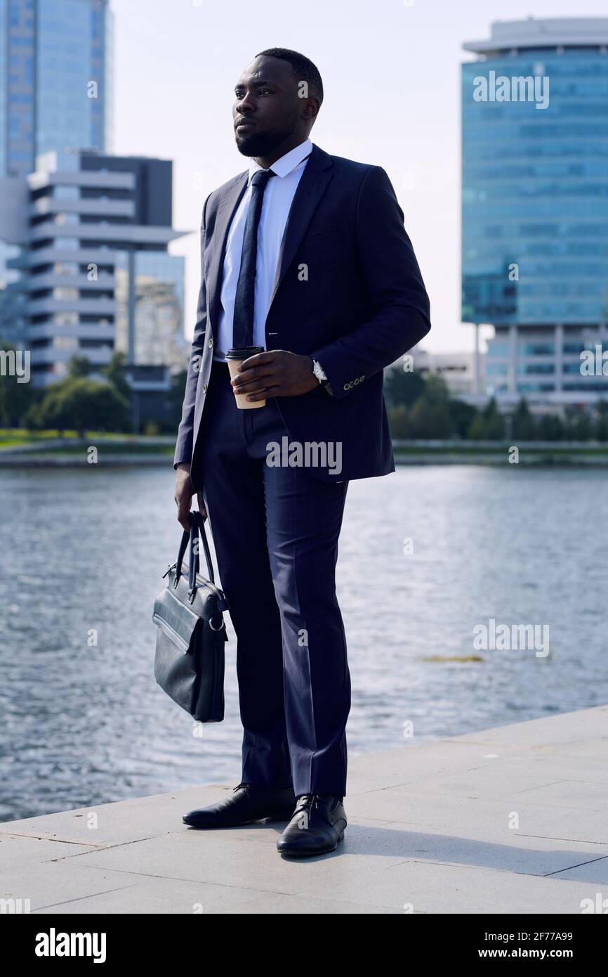 Junger erfolgreicher afrikanischer Geschäftsmann mit Handtasche, die am  Flussufer trinkt Während Sie an sonnigen Tagen im städtischen Umfeld stehen  Stockfotografie - Alamy