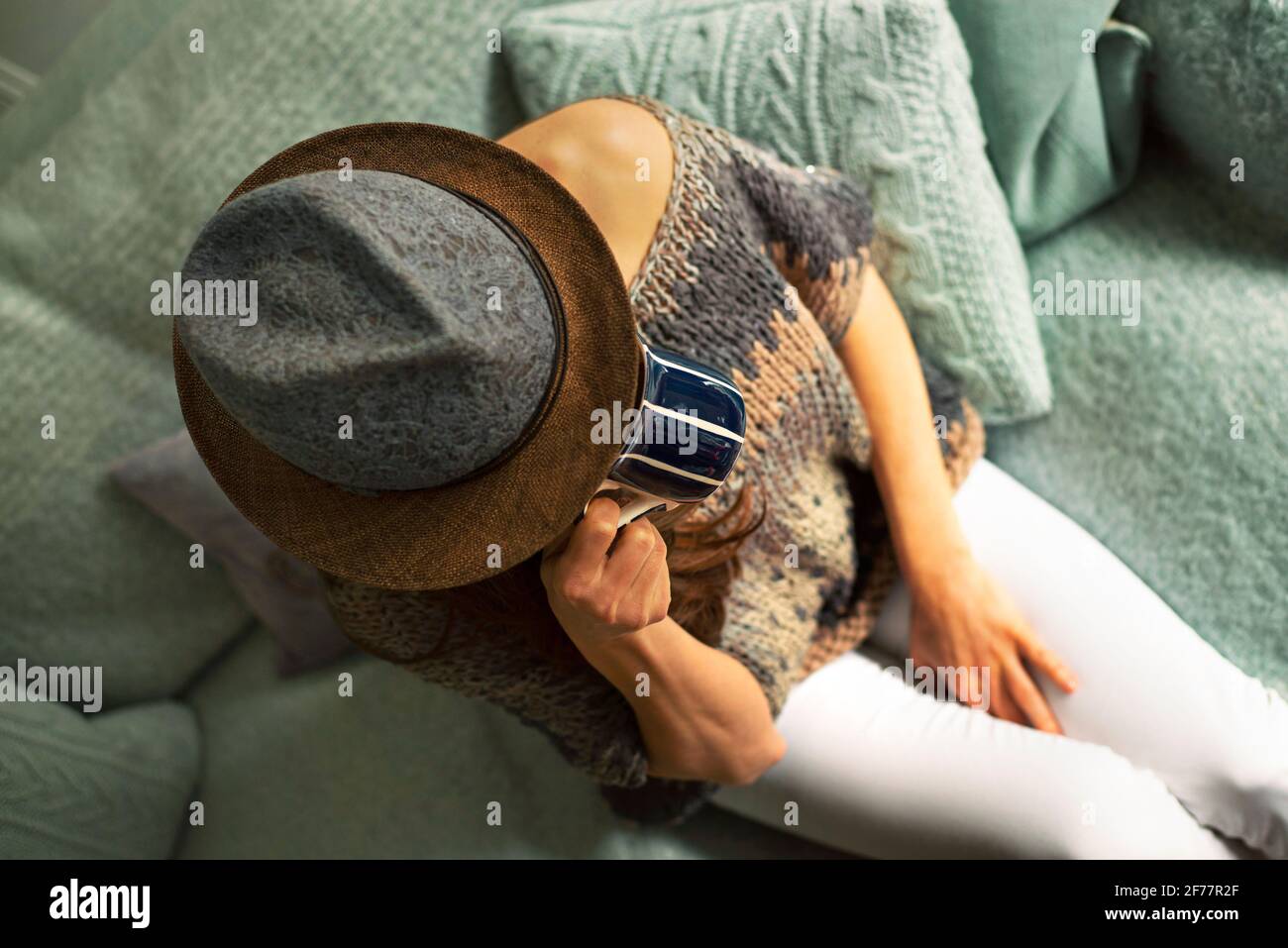 Von oben gesehen, wie eine Frau in stilvoller Strickmode und Hut auf einem gemütlichen Enteneier-Sofa sitzt und ein heißes Getränk trinkt. Indoor Lifestyle, Tee oder Kaffeepause Konzept Stockfoto