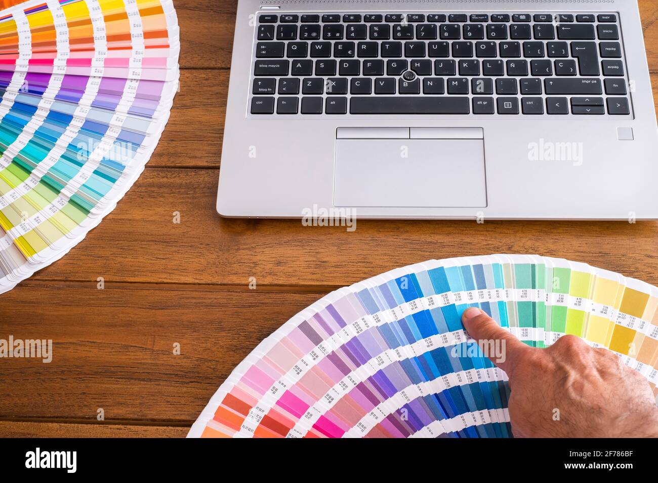 Eine Hand mit einem Finger, die eine Farbe auf einem offenen Farbfeld auswählt, wobei alle verschiedenen Farben auf einem Holztisch mit einem Laptop horizontal ausgeklappt sind Stockfoto