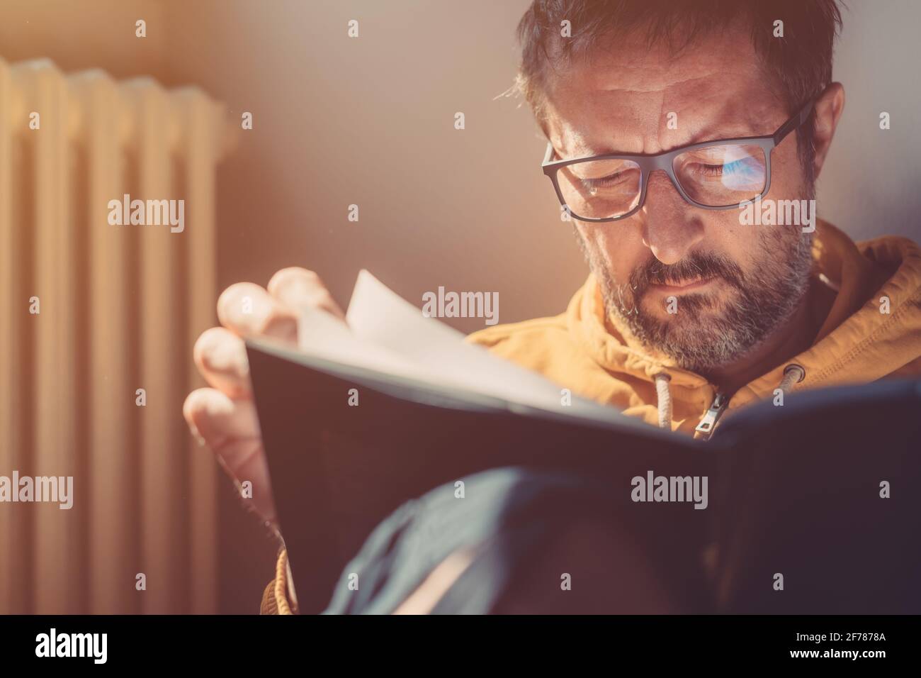 Nachdenklicher Mann mittleren Erwachsenen, der zu Hause ein Buch liest, Nahaufnahme-Porträt mit Kopfschuss und selektivem Fokus, Sonnenlicht, das vom Fenster scheint Stockfoto