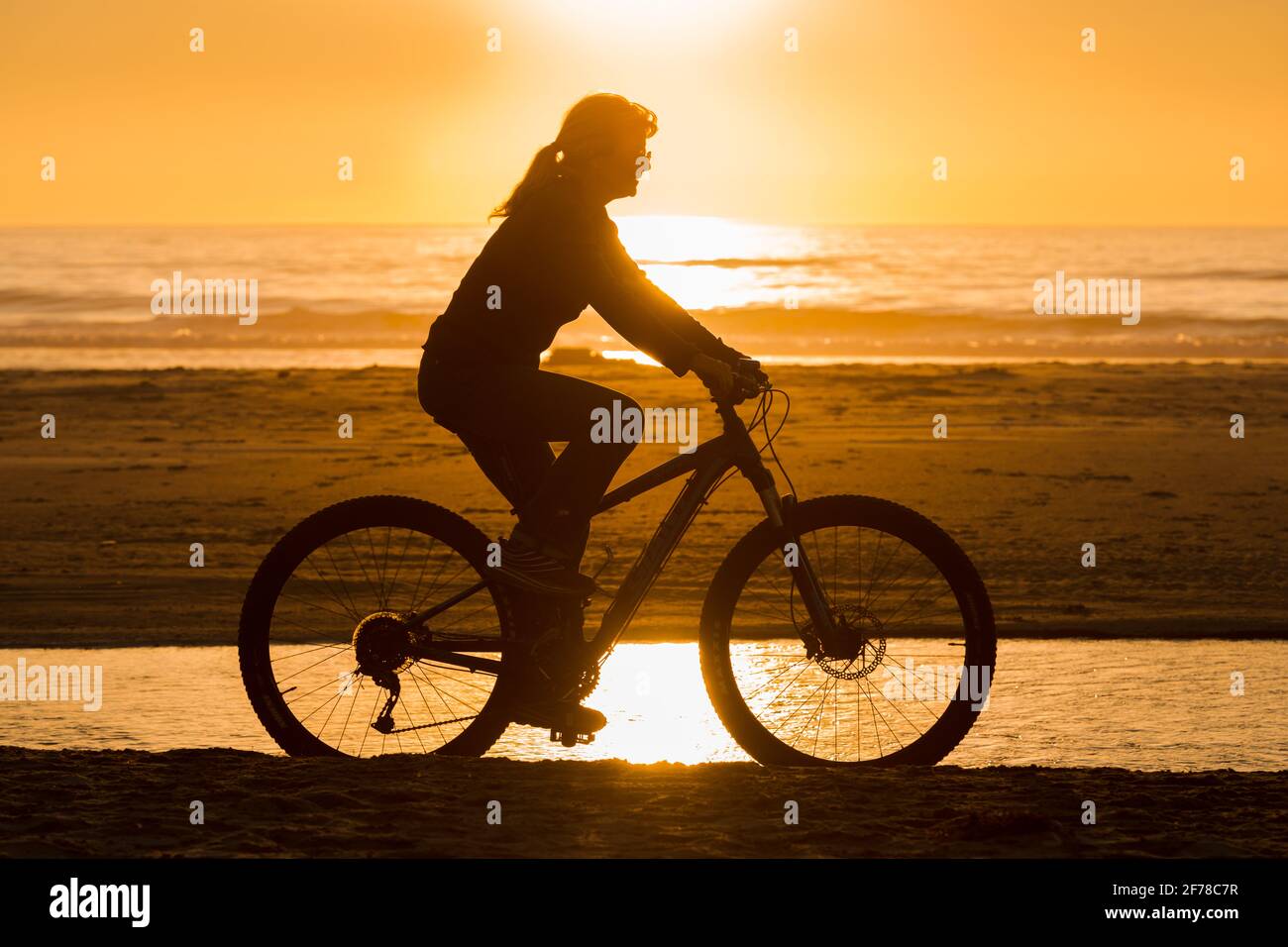Eine Frau fährt am Strand mit dem Fahrrad Oregon Küste gegen eine orange untergehende Sonne als Teil Eines gesunden Lebensstils Stockfoto