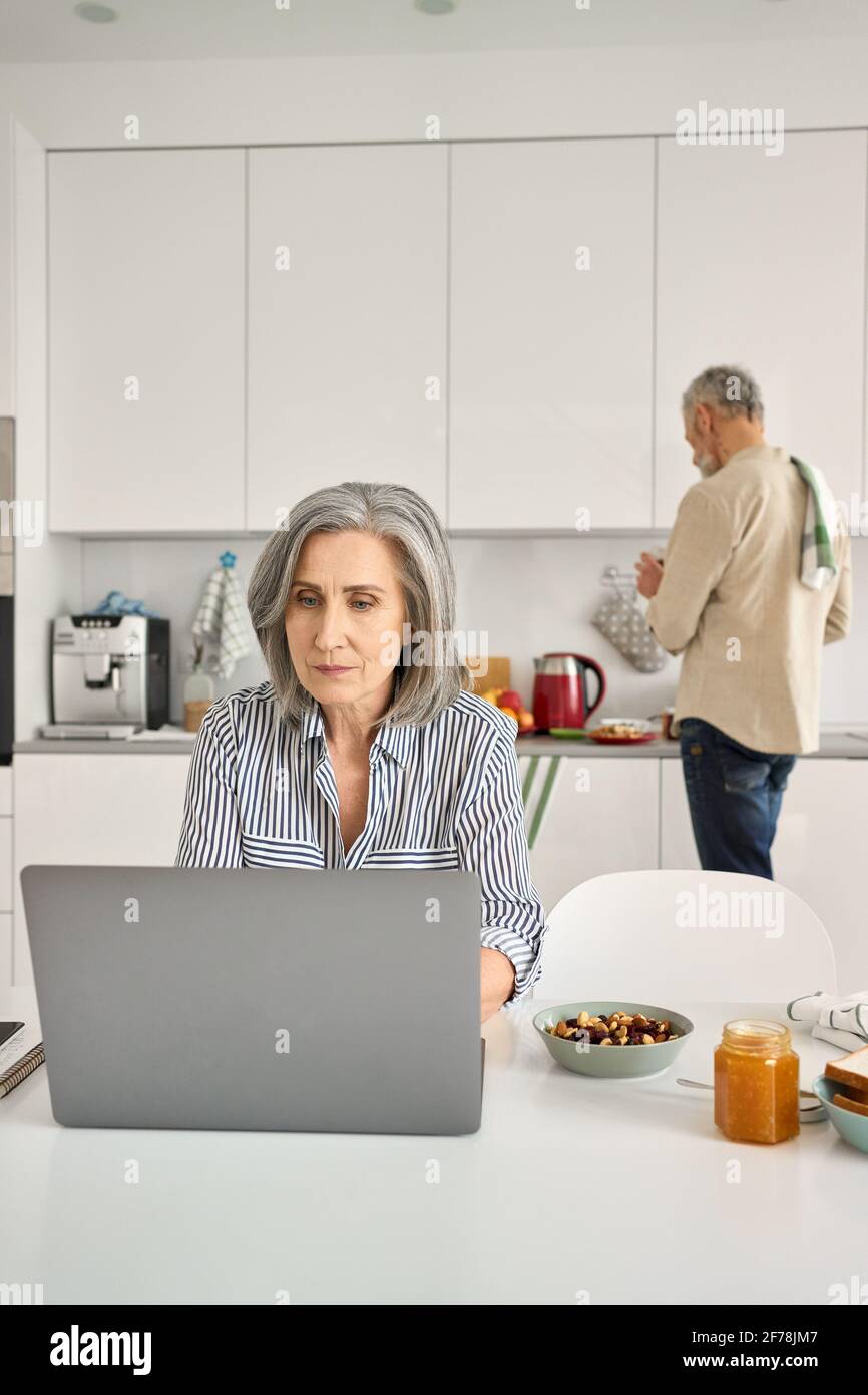 Reife Frau arbeitet online im Home Office, während alter Mann in der Küche kocht. Stockfoto