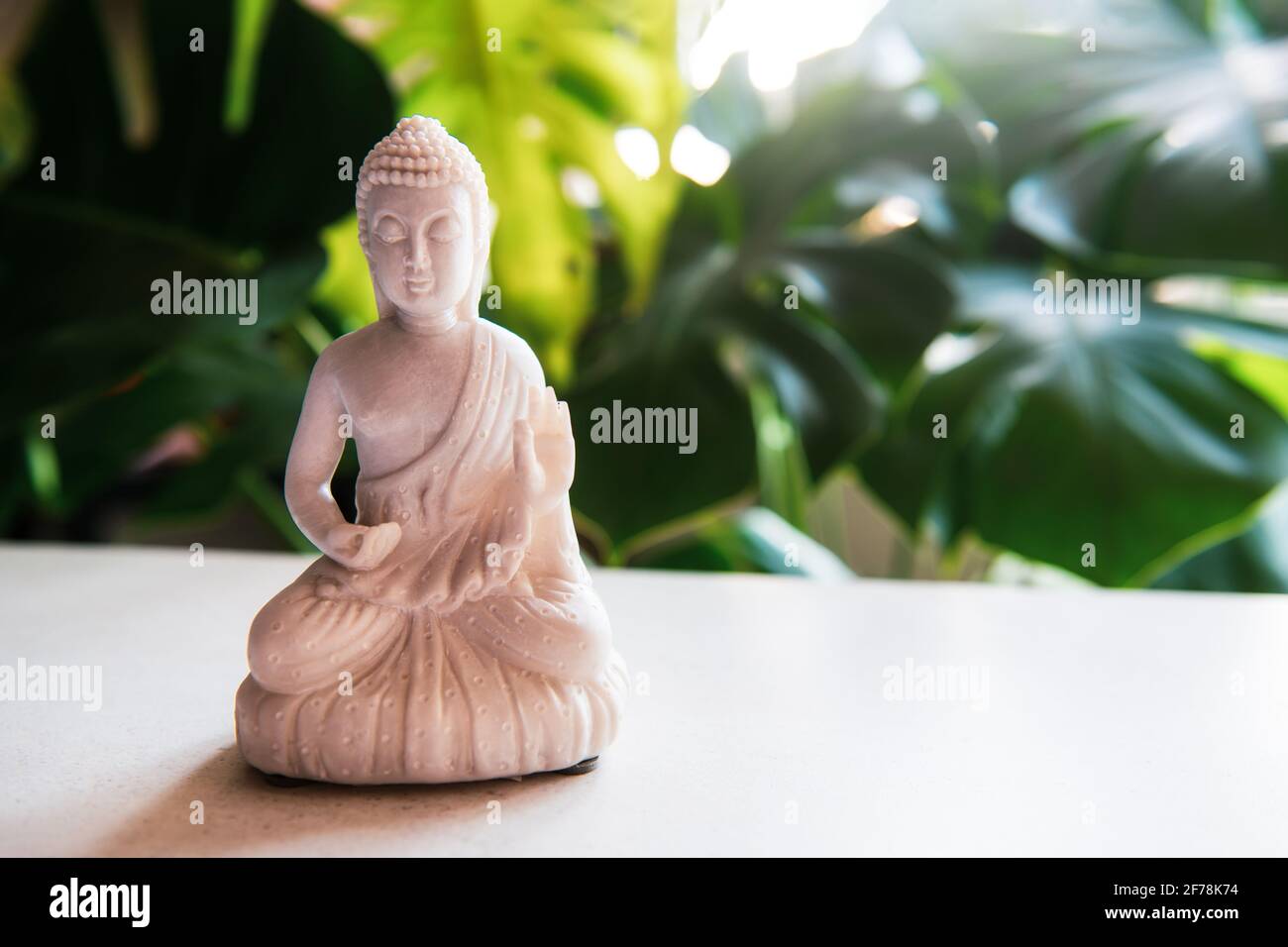 Dekorative weiße Buddha-Statue mit grüner Monstera-Pflanze auf dem Hintergrund. Meditations- und Entspannungsritual. Exotische Massage. Selektiver Fokus. Kopieren Stockfoto