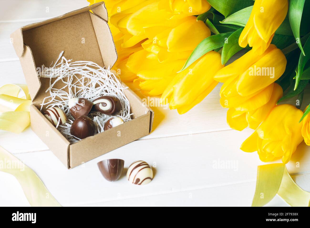 Schokolade Bonbons in Bastelkiste und Bouquet von gelben Tulpen Auf weißer Holzoberfläche Stockfoto