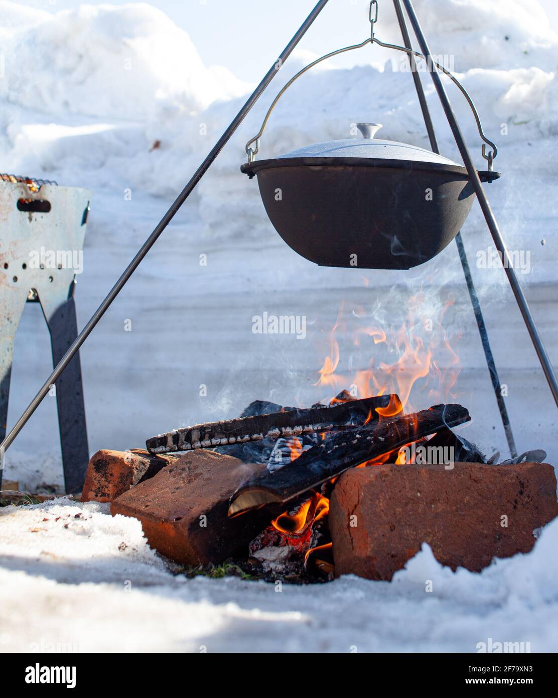 Über dem Feuer hängt ein Topf, in dem man Essen kochen kann. An einem Haken auf einem Stativ tritt Dampf aus der Pfanne aus. Winter Camping Kochen im Freien Stockfoto