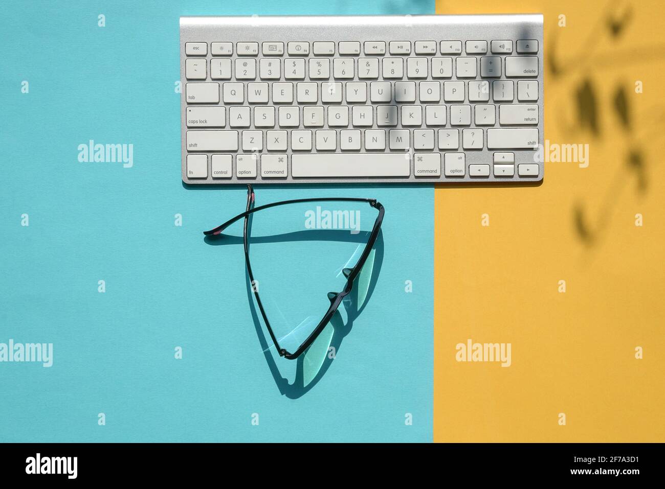 Weiße Tastatur und Lesebrille auf blauem und gelbem Hintergrund oben. Mit abstrakten Schatten der Blätter auf dem Tisch. Stockfoto