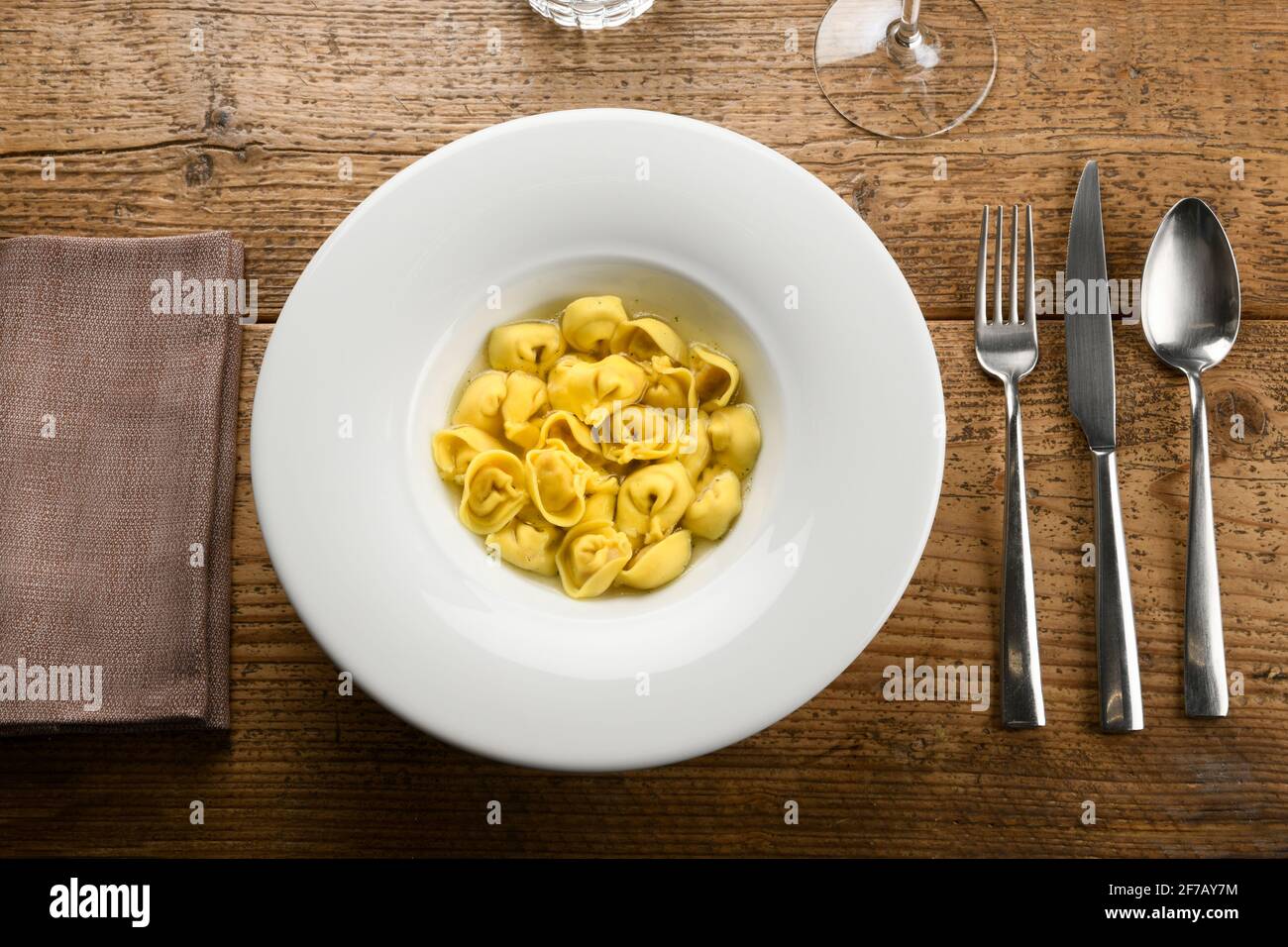 Draufsicht auf eine Schüssel mit köstlicher Cappelletti-Pasta mit Brühe Platziert auf Holztisch neben Serviette und Besteck Stockfoto