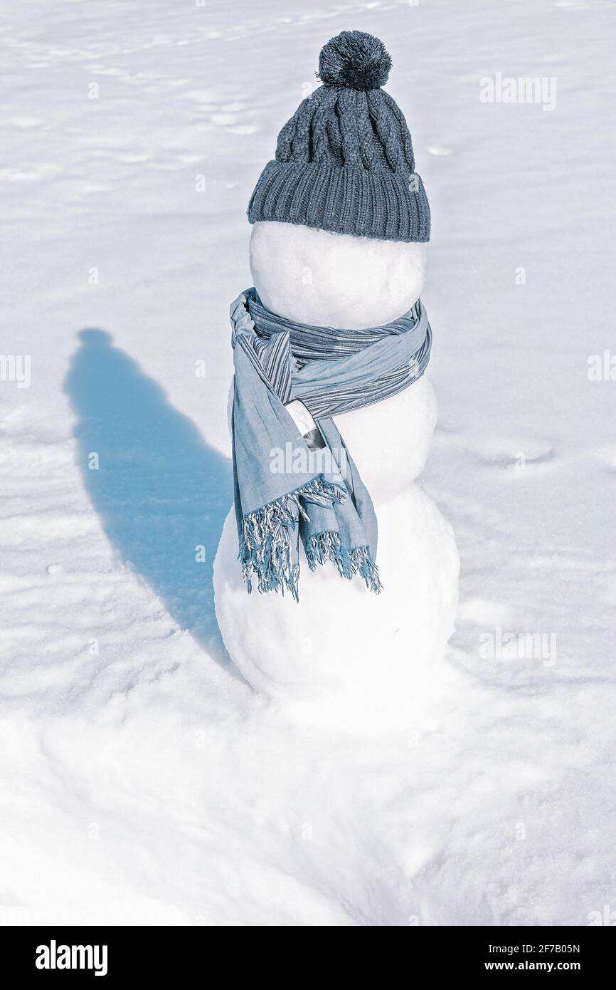 Ein Schneemann mit blauem Hut und Schal steht im weißen Feld. Ein Eismann auf einem hellen auffälligen Hintergrund. Sonniger Tag, eiskalt, Nahaufnahme. Stockfoto