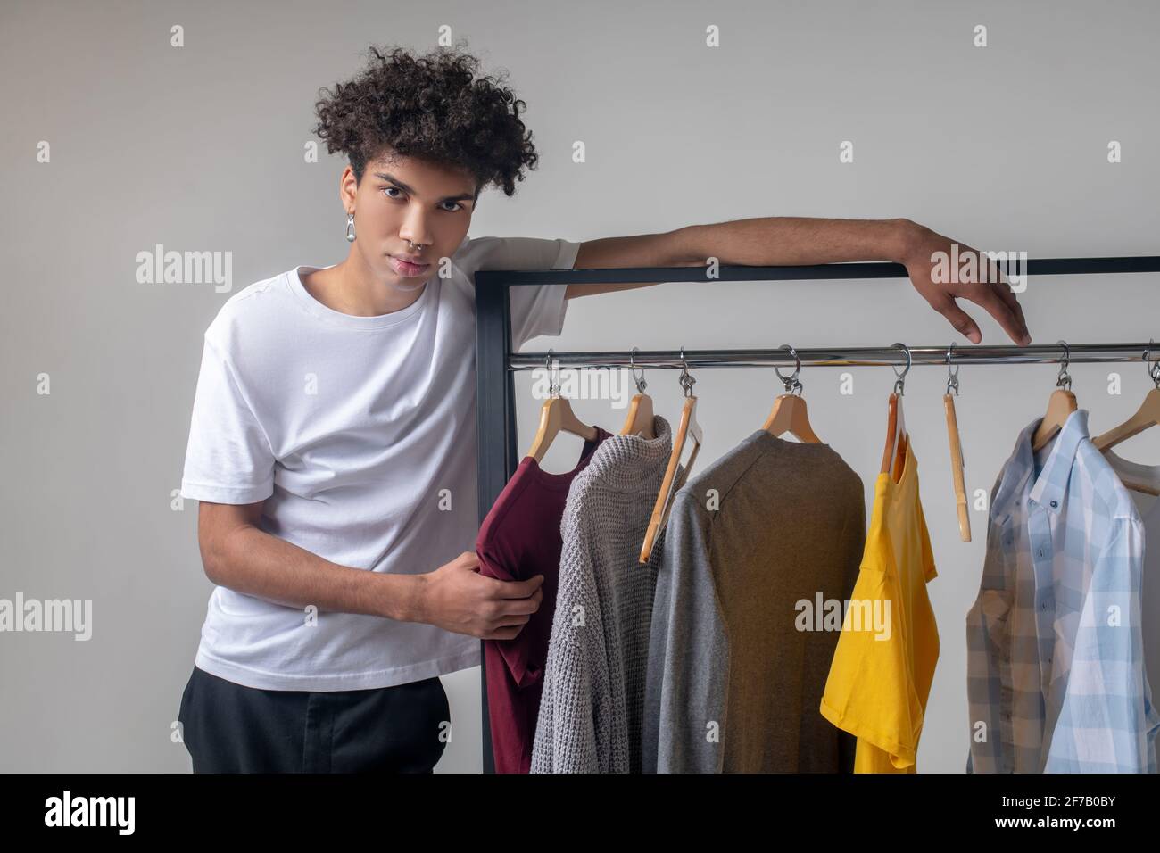 Lockiger junger Mann, der neben den Kleiderbügeln mit Kleidung steht Stockfoto