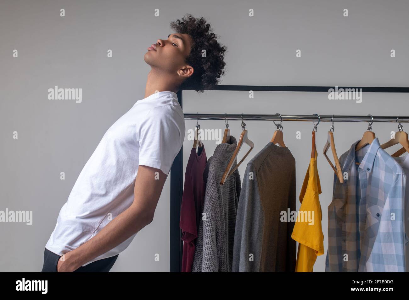 Lockiger junger Mann, der neben den Kleiderbügeln mit Kleidung und steht Sieht nachdenklich aus Stockfoto