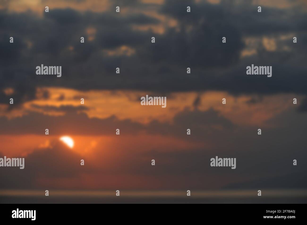 Bokeh-Effekt von Ozean und Himmel bei Sonnenuntergang mit dunklen Wolken und teilweise verborgener Sonne, die eine launische, nostalgische Atmosphäre und einen verschwommenen Hintergrund schaffen Stockfoto