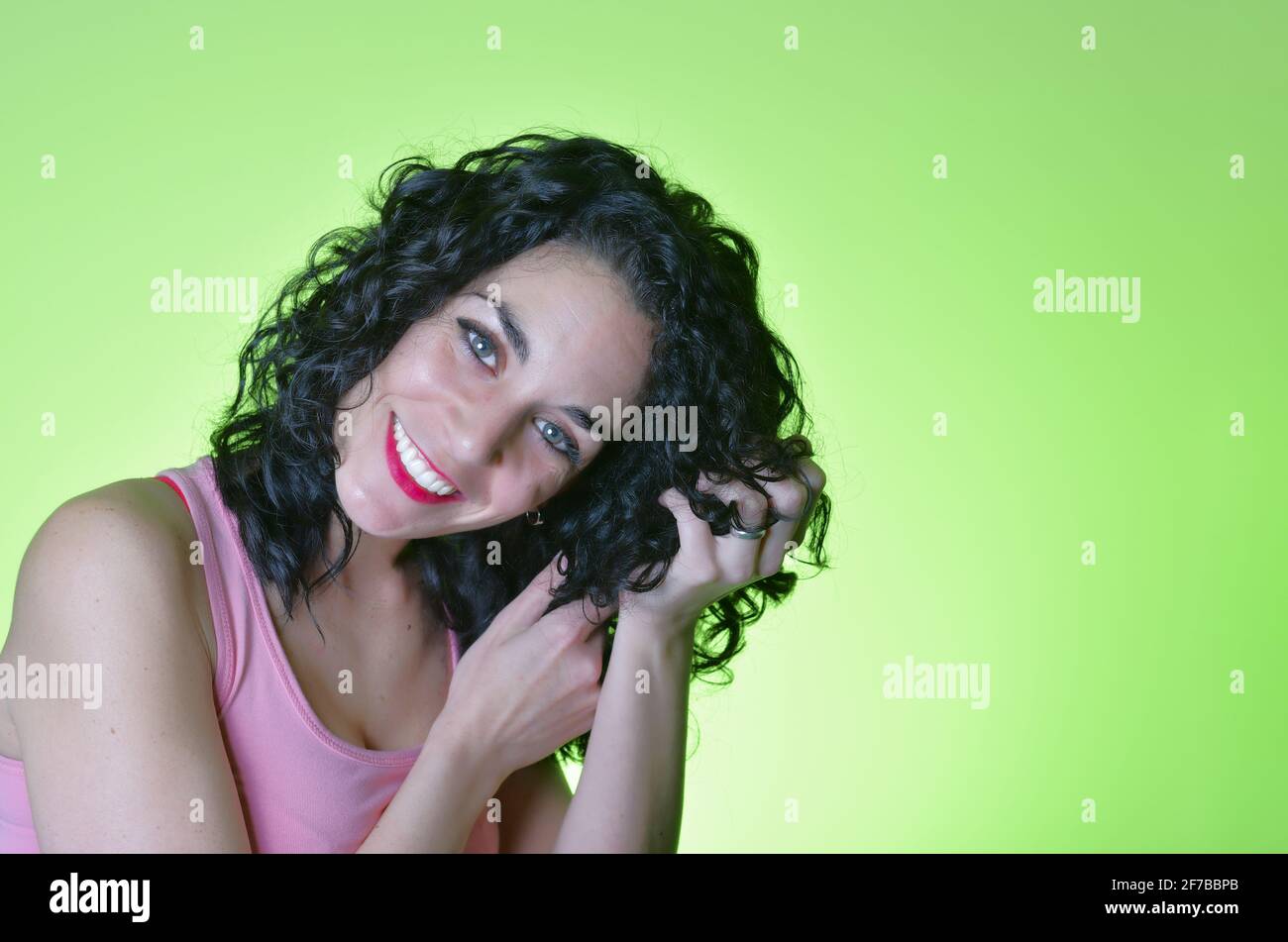 Junge Frau mit lockigen schwarzen Haaren und grünen Augen, die sich um ihr lockiges Haar kümmert und der lockigen Mädchenmethode folgt. Haarpflegekonzept. Grüner Backgroun Stockfoto