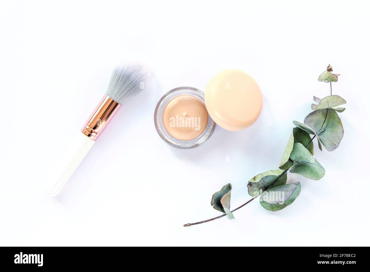 Concealer, Make-up Pinsel und grüner Eukalyptuszweig auf weißem Hintergrund. Draufsicht, flach liegend. Konzept der Naturkosmetik Stockfoto