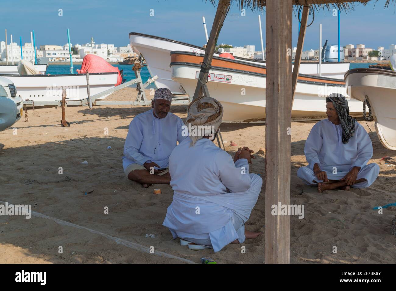 Drei Fischer, die sich im Schatten eines Baldachins zwischen ihren Fischerbooten am Strand von Sur, Oman, treffen. Stockfoto