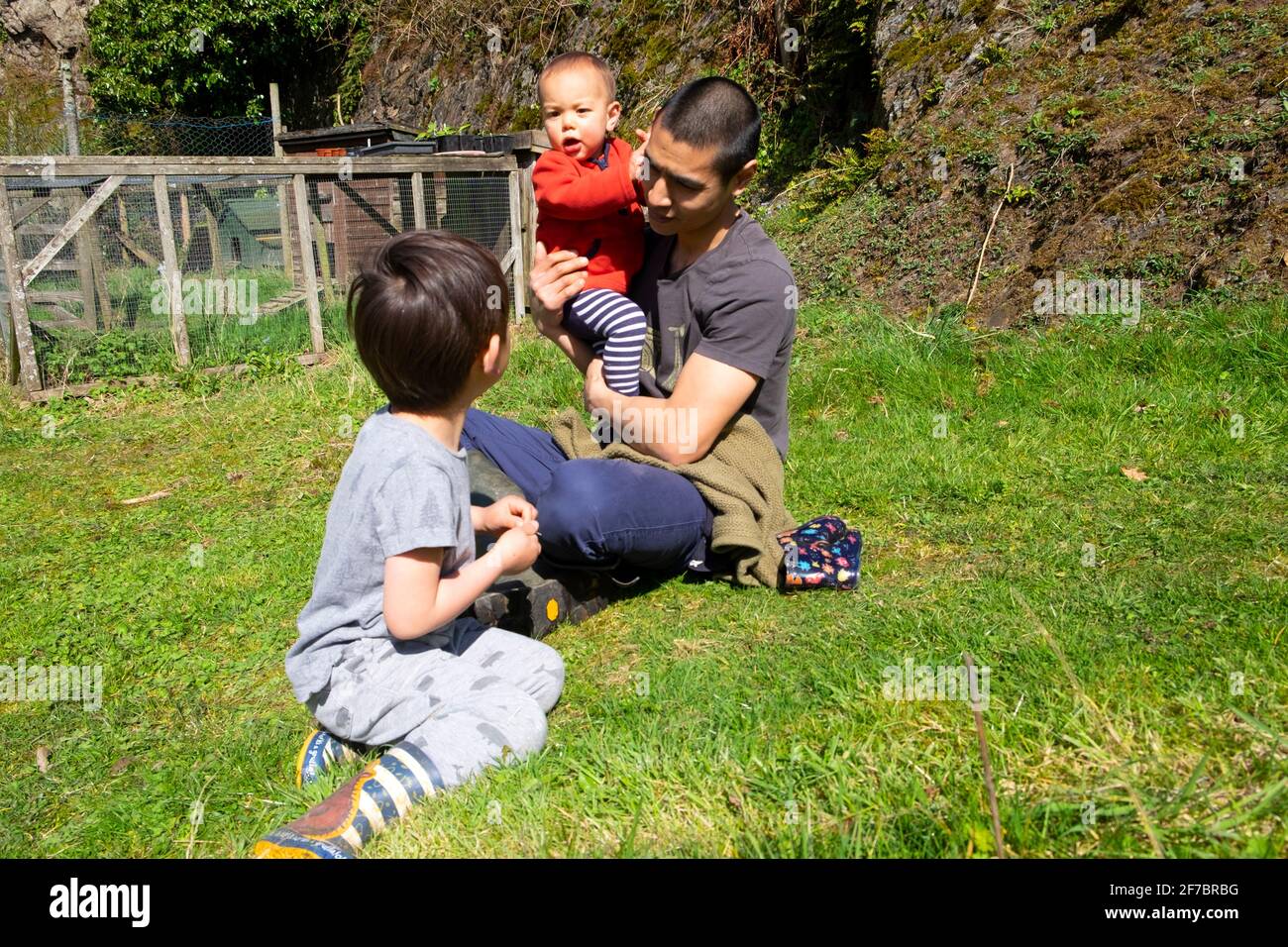 Asiatische Mann Vater Vater mit zwei junge Kinder Jungen Bruder Beobachten Sie, wie Sie im Frühling in Carmarthenshire Wales ein Kleinkind im Freien halten UK KATHY DEWITT Stockfoto