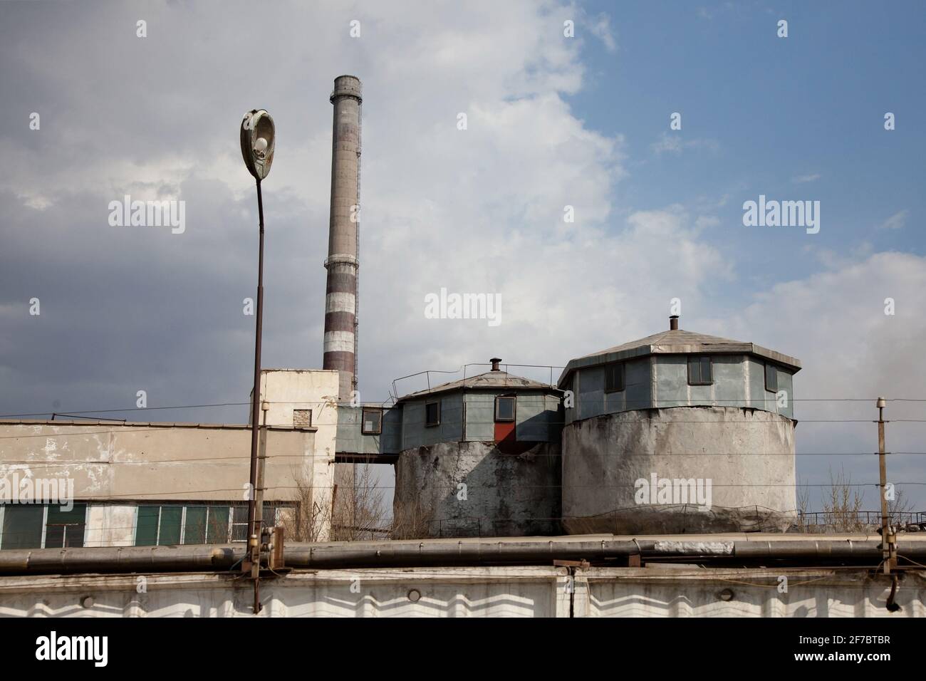 Veraltete sowjetische Bergbau- und Verarbeitungsfabrik. Grauer Rauchstapel aus Beton und zwei runde Silos. Blauer Himmel mit grauen Wolken. Stockfoto