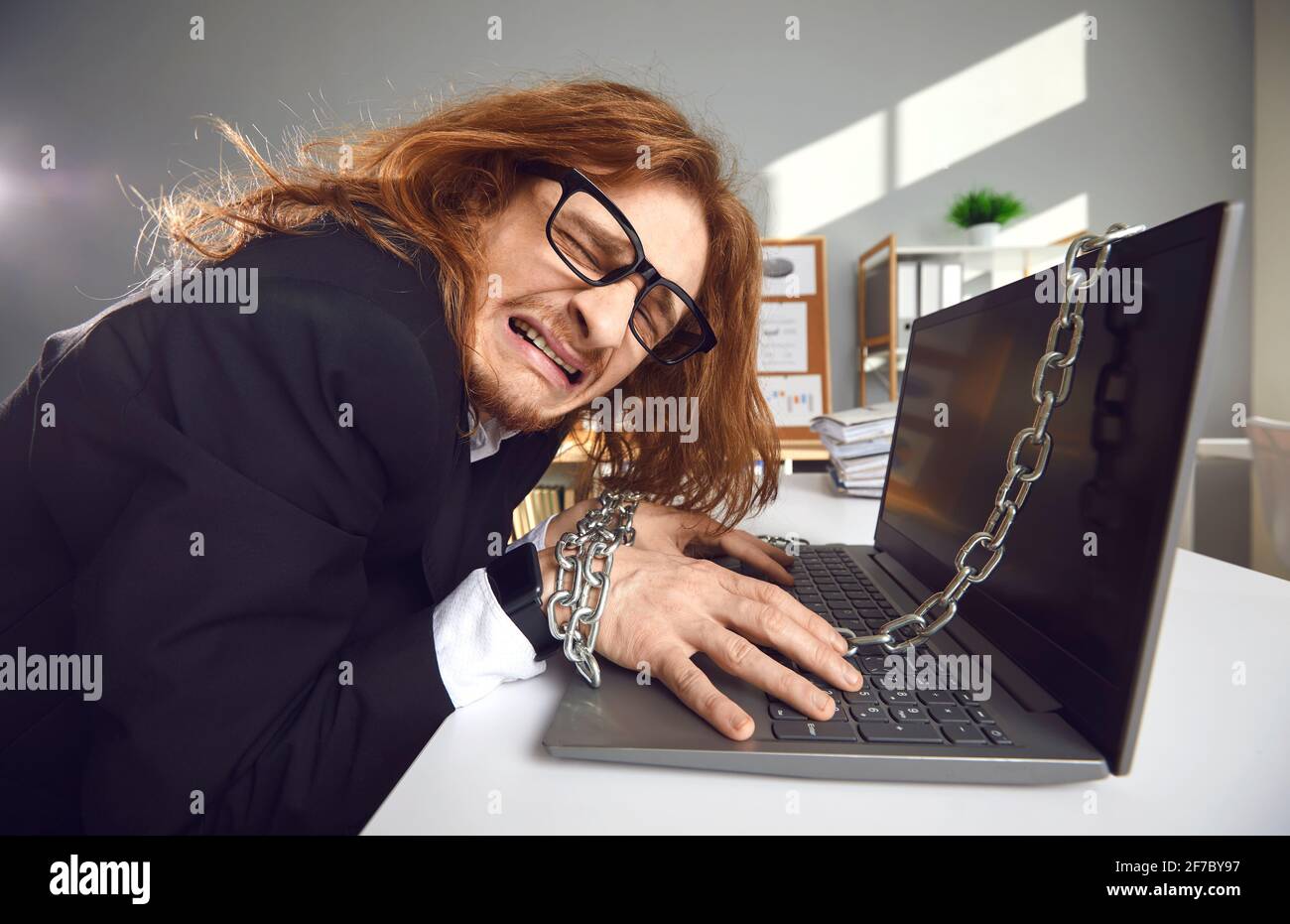 Seitenansicht eines müden, verrückten Büroarbeiters, der Überstunden an einen Laptop gekettet arbeitet und weint. Stockfoto