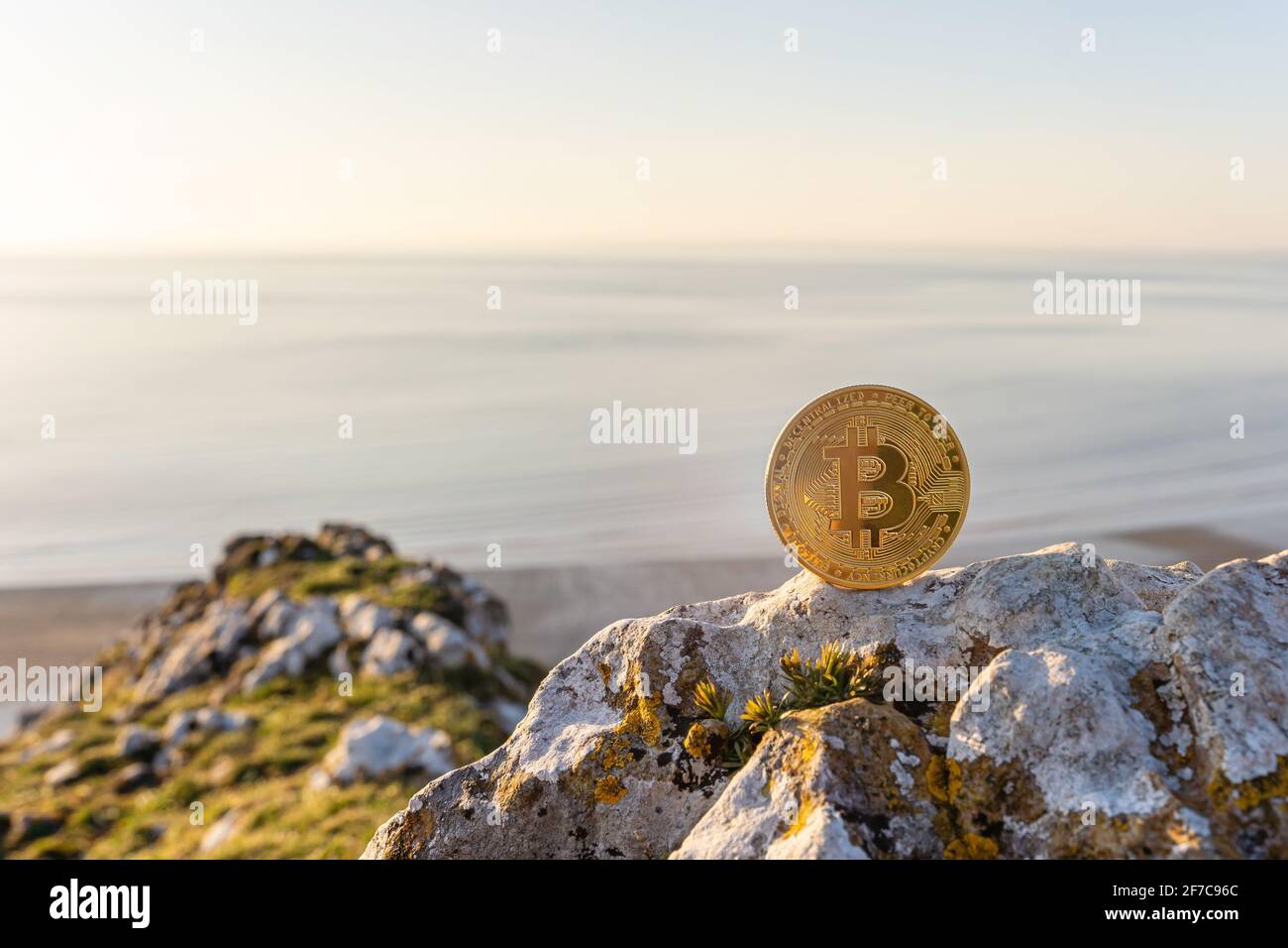 Nahaufnahme von Bitcoin auf einem Stein im Freien mit schönem Meer bei Sonnenuntergang Hintergrund mit Kopierraum. Einzelne Gold glänzende BTC Kryptowährung Münze. Stockfoto