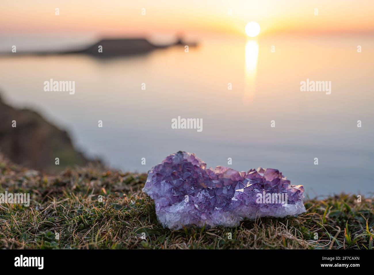 Amethyst Kristall Druse liegt auf Gras mit Meer bei Sonnenuntergang Hintergrund mit Kopieplatz. Einzelne rohe, natürliche violette Geode im Freien Stockfoto
