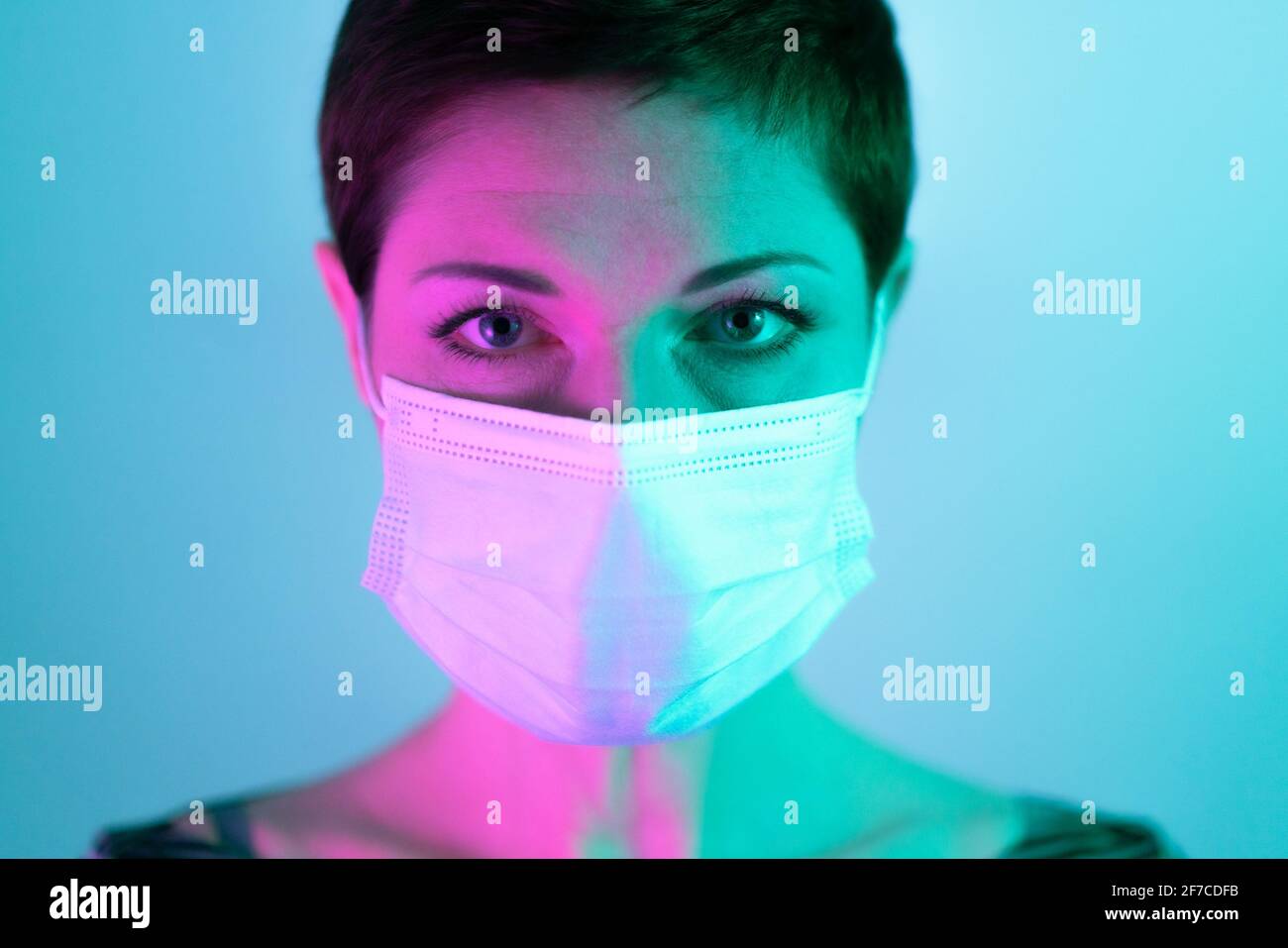Schöne Frau mit kurzen Haaren, die eine Gesichtsmaske trägt und besorgt und deprimiert in violettem und grünem Licht aussieht. Leben in Quarantäne. Umgang mit coro Stockfoto