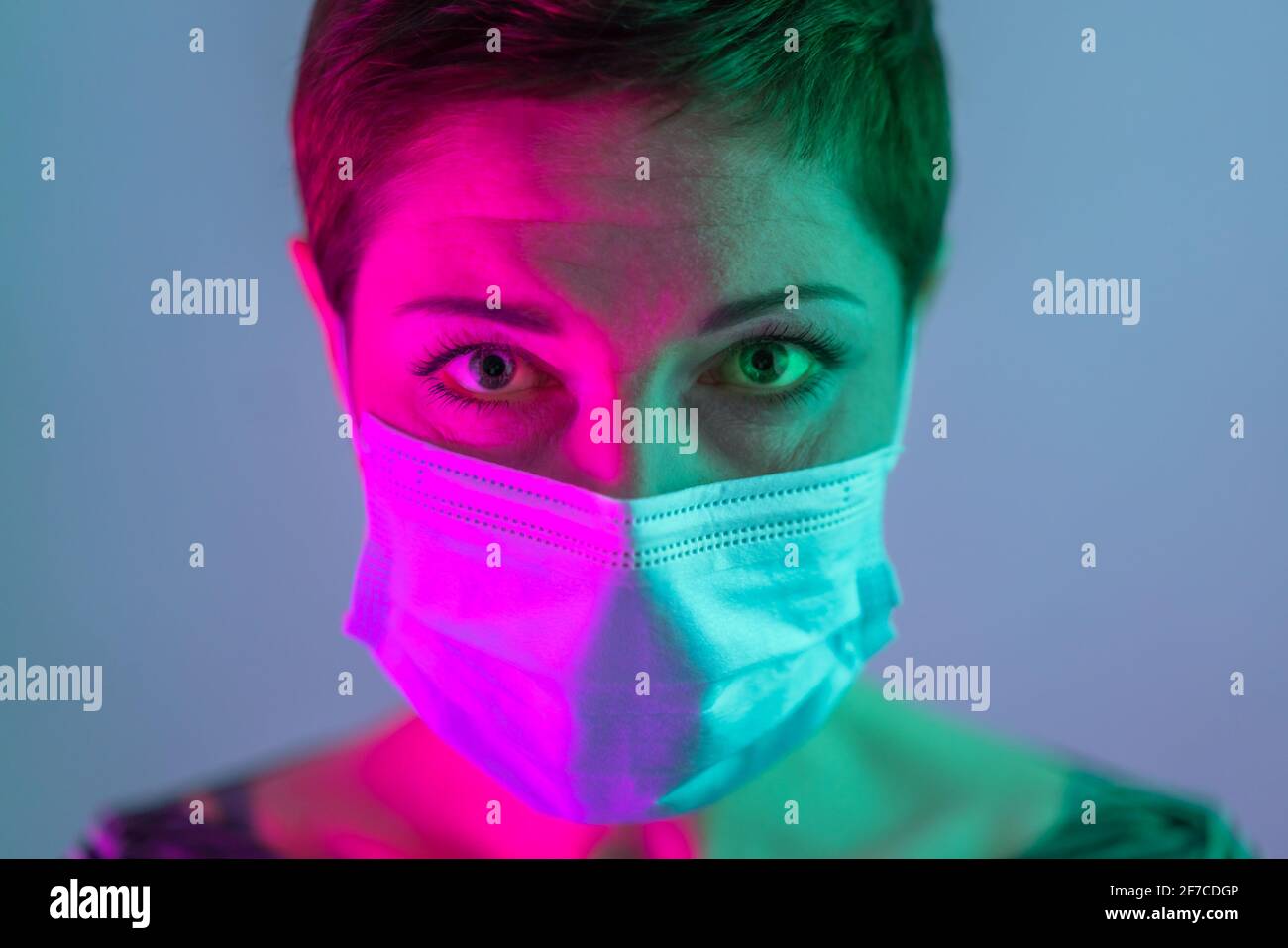Schöne Frau mit kurzen Haaren, die eine Gesichtsmaske trägt und besorgt und deprimiert in violettem und grünem Licht aussieht. Leben in Quarantäne. Umgang mit coro Stockfoto
