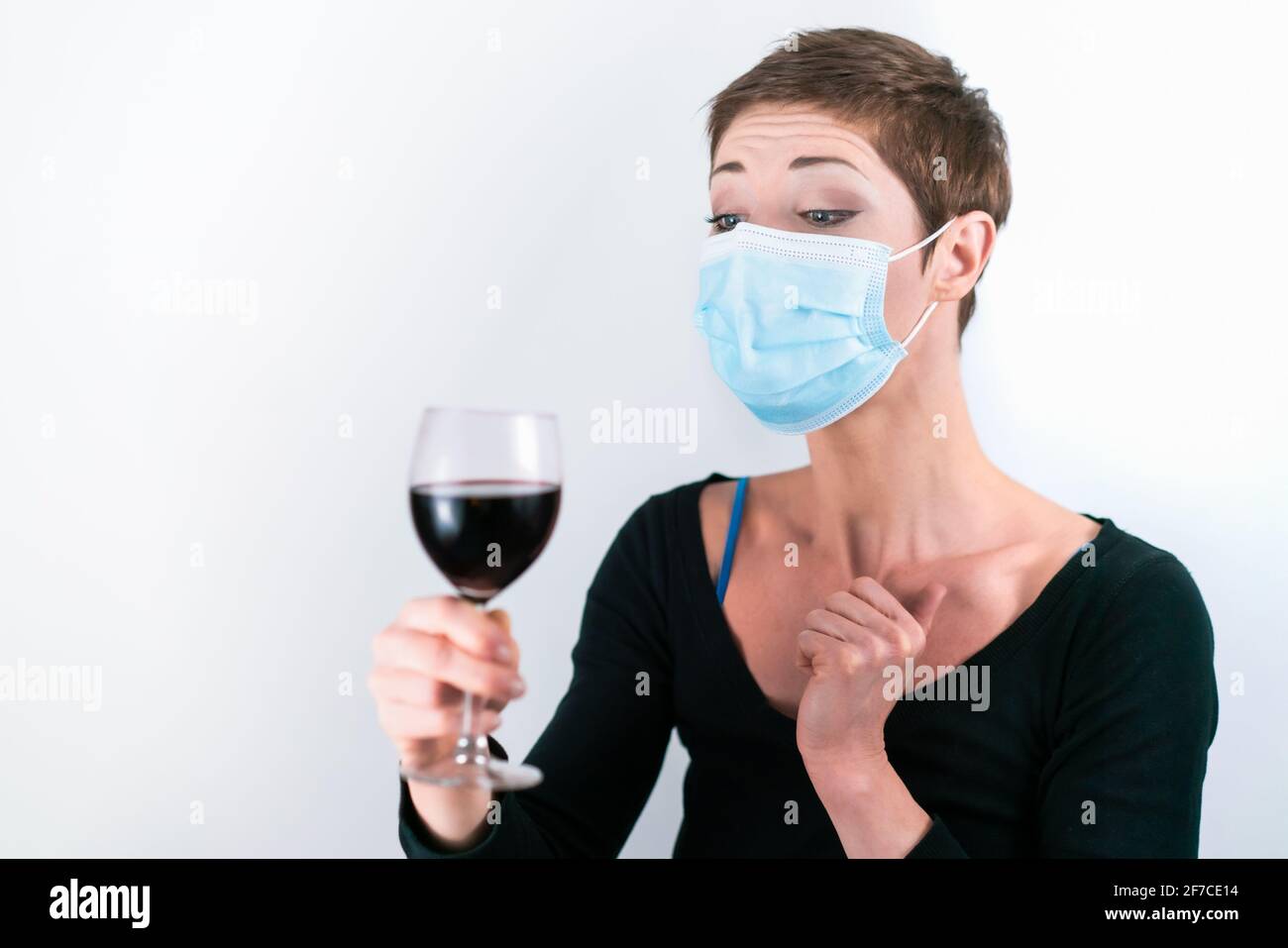 Schöne Frau mit kurzen Haaren, die eine Gesichtsmaske trägt und versucht, einen Weg zu finden, wie man aus Rotwein aus einem Glas trinkt. Leben in Quarantäne. Umgang mit Cor Stockfoto