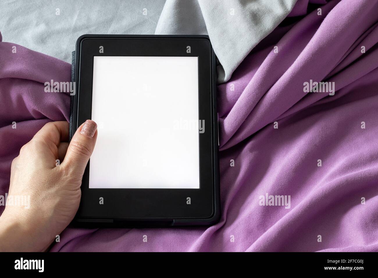 Ein modernes elektronisches E-Reader mit einem leeren Bildschirm in weiblicher Hand auf einem grauen und violetten Bett. Mockup Tablet auf Microfaser-Bettwäsche Nahaufnahme Stockfoto