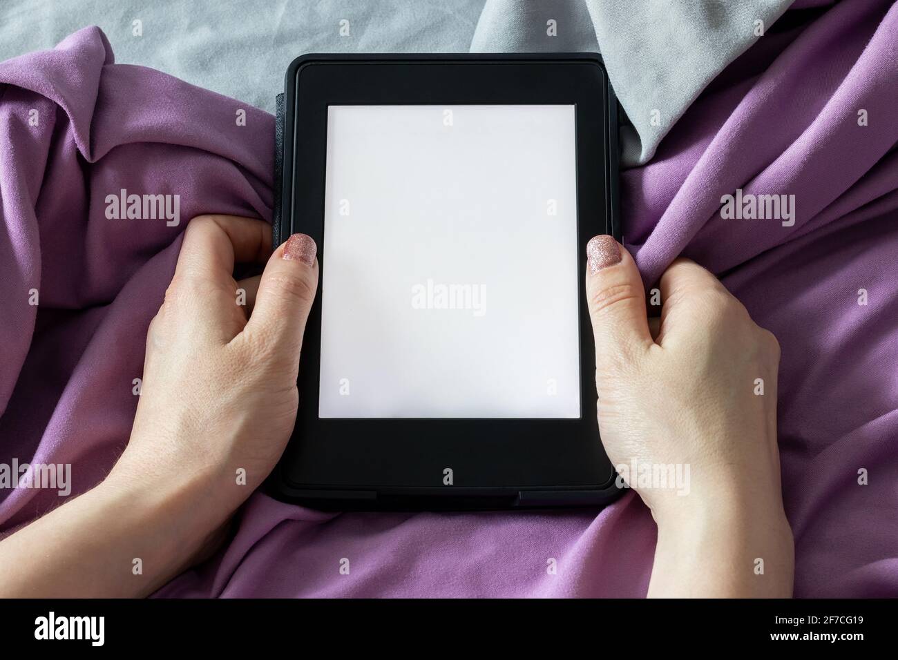 Ein modernes elektronisches E-Reader mit einem leeren Bildschirm in weiblichen Händen auf einem grauen und violetten Bett. Mockup Tablet auf Microfaser-Bettwäsche Nahaufnahme Stockfoto