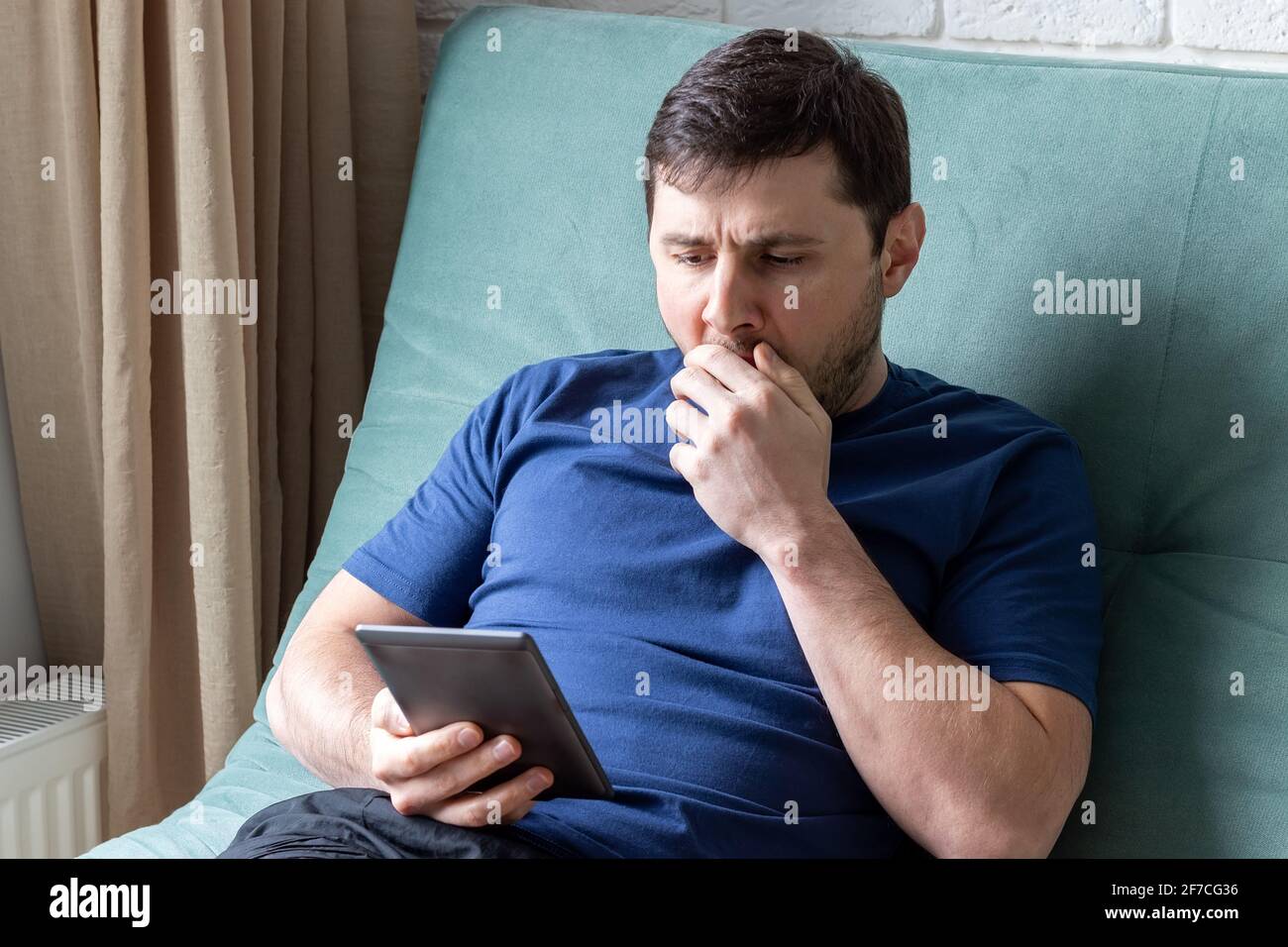 Der Mensch gähnt beim Lesen eines E-Books. Ein ruhender Mann in einem blauen T-Shirt sitzt auf einem grünen Sofa mit einem elektronischen Buch in der einen Hand, gähnt Stockfoto