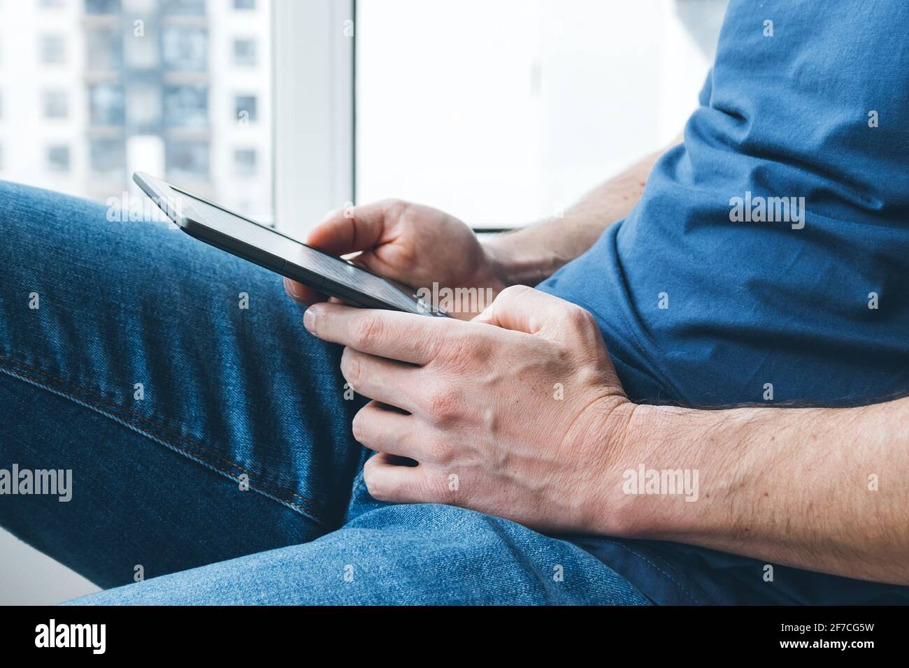 Ein Mann in einem blauen T-Shirt und Jeans hält ein elektronisches E-Reader-Buch in den Händen. Bücher zu Hause am Fenster lesen. Selektiver Fokus Stockfoto