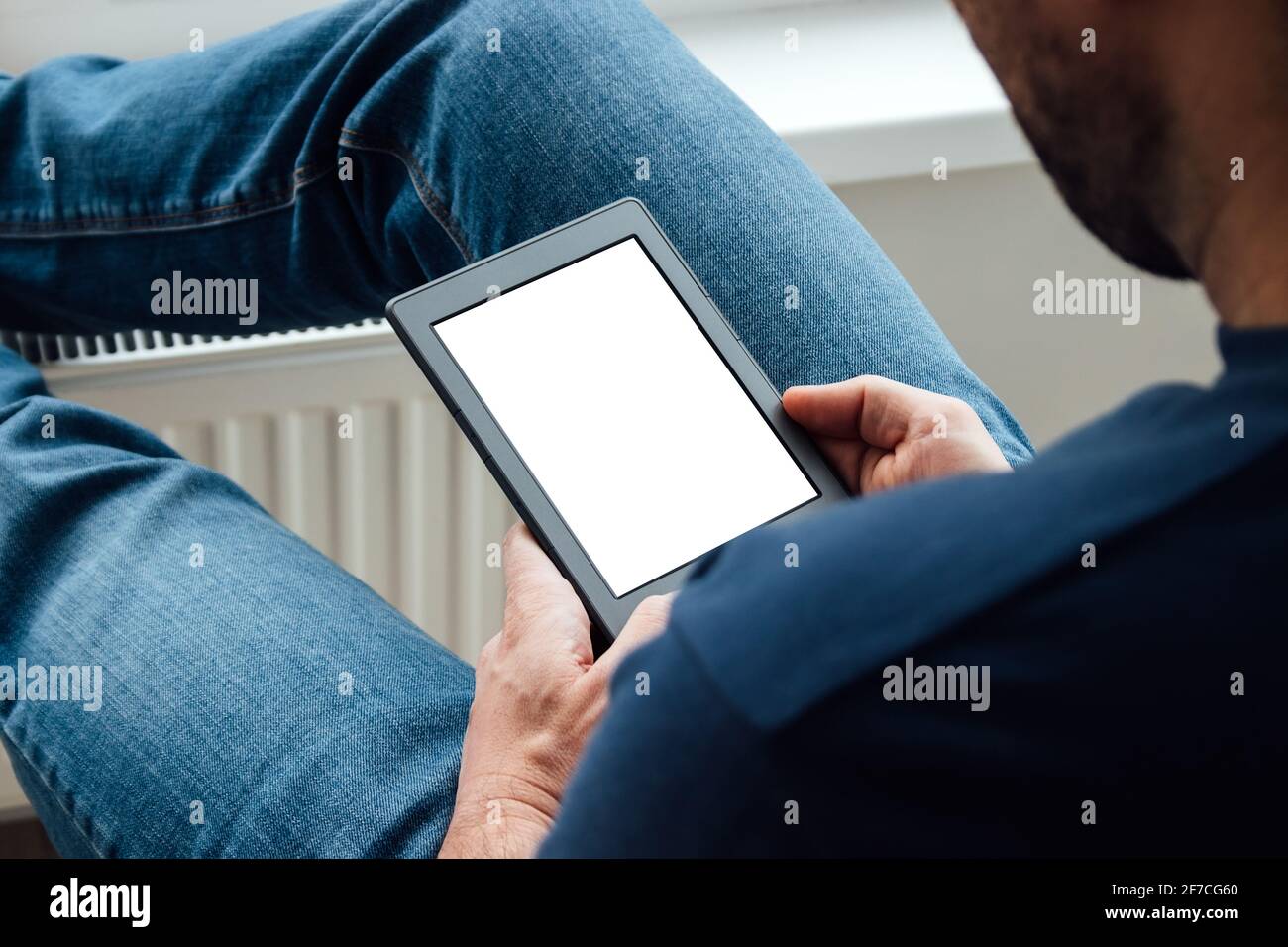 Ein Mann in einem blauen T-Shirt und Jeans hält ein elektronisches E-Reader-Buch mit weißem, leerem Mockup-Bildschirm in den Händen. Bücher zu Hause am Fenster lesen. Stockfoto