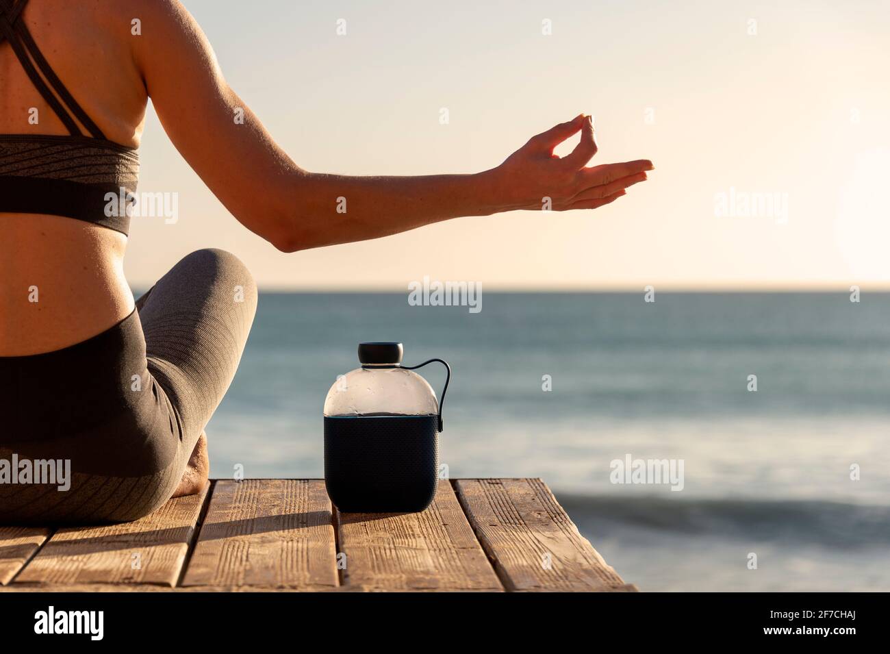 Nahaufnahme einer Frau, die bei Sonnenaufgang oder Sonnenuntergang am Meer meditiert, mit einer Flasche Wasser, um hydratisiert zu bleiben. Stockfoto