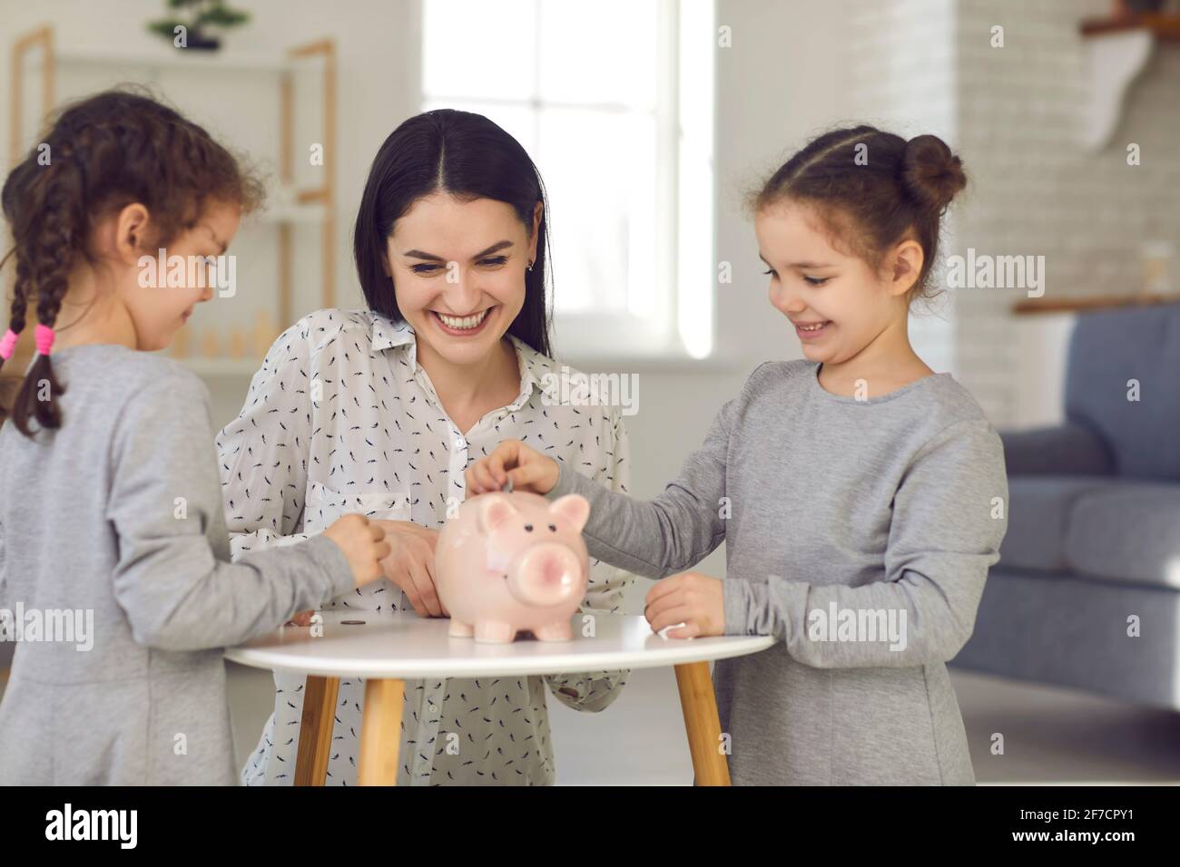 Glücklich lächelnd kleine niedliche Töchter in grauen Kleidern und jung Mutter legt Münzen auf das Sparschwein Stockfoto