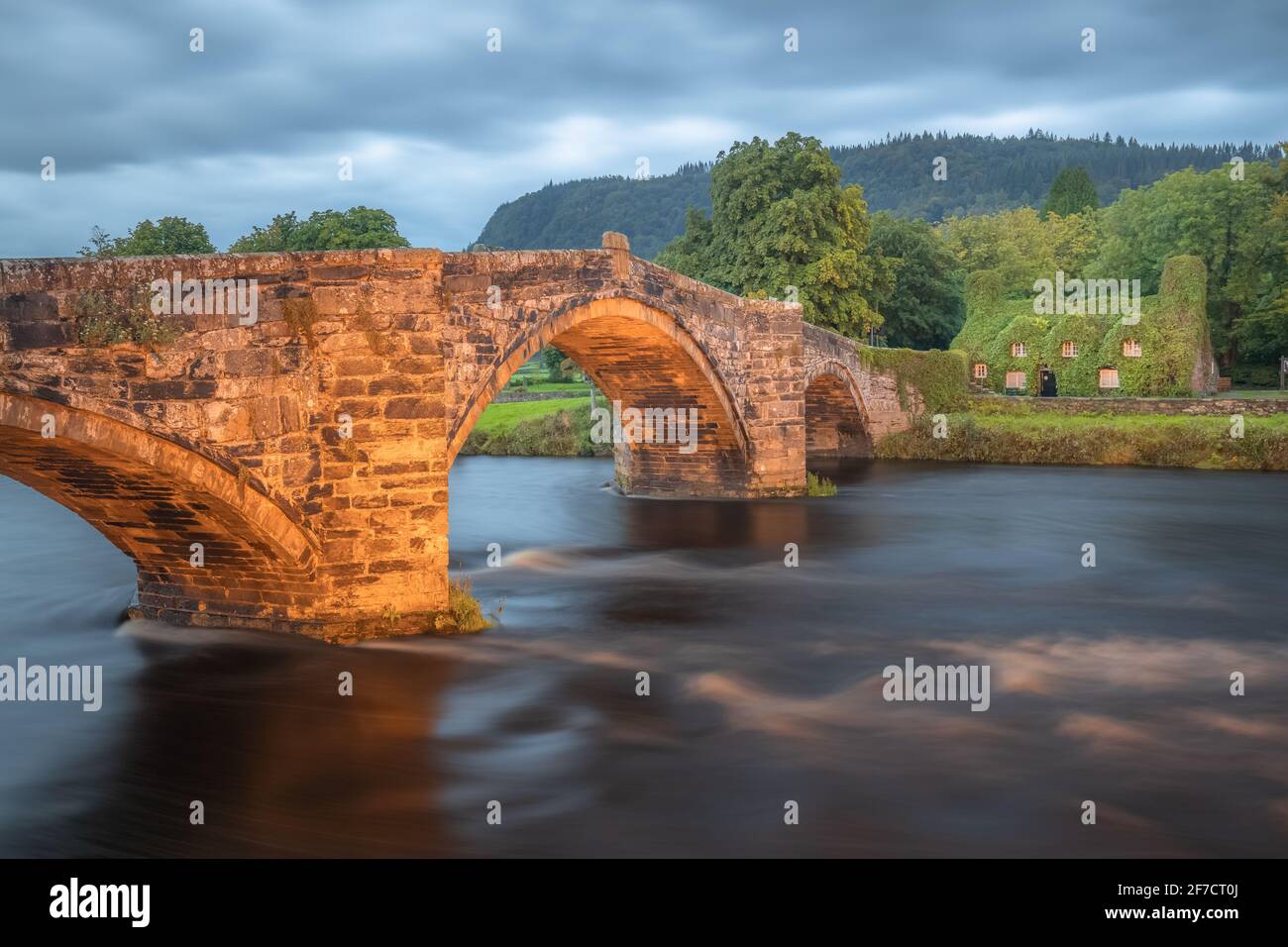 Alte römische Brücke Pont Fawr über den River Conwy in Richtung eines mit Efeu bedeckten Steinhauses aus dem 15. Jahrhundert im walisischen Dorf Llawnrst, Nordwales Stockfoto