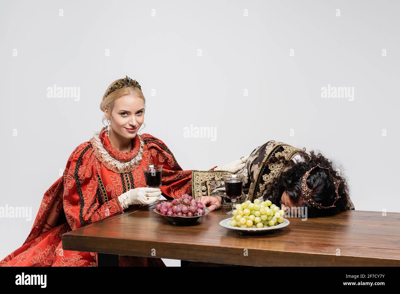 Glückliche Königin hält ein Glas Rotwein in der Nähe von vergifteten hispanischen König in mittelalterlicher Kleidung isoliert auf Weiß Stockfoto