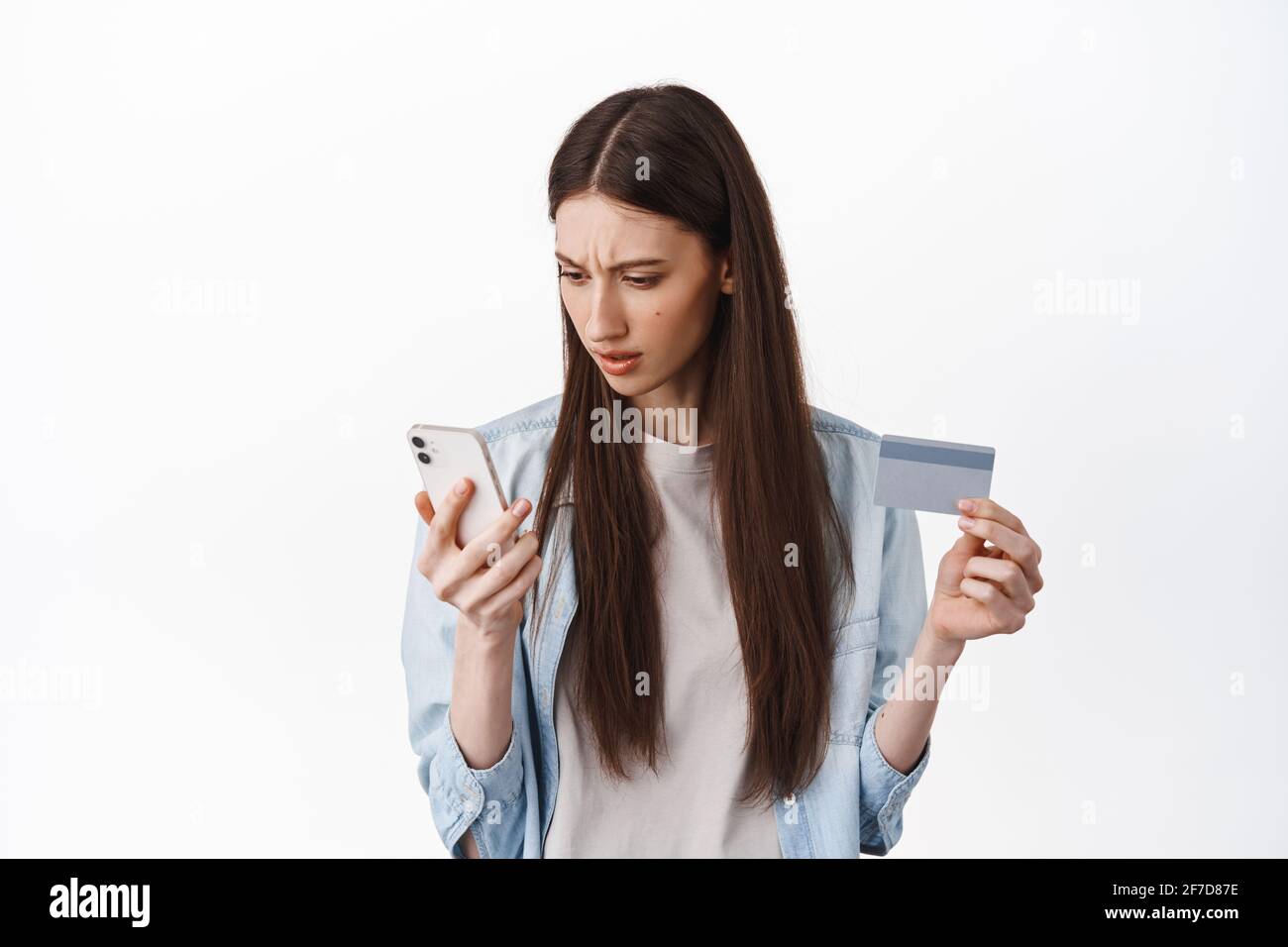 Online-Shopping. Brunette Mädchen sieht verwirrt auf Smartphone-Bildschirm, hält Kreditkarte, kann nicht verstehen, wie Karte registrieren, um eine Bestellung aufzugeben, stehend Stockfoto