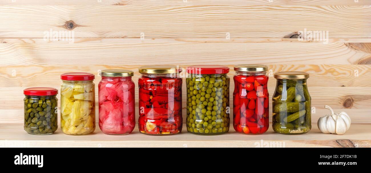 Gemüsekonserven auf einem Holzregal. Kapern, Artischocke, Ingwer, getrocknete Tomaten, grüne Erbsen, Baby rote Paprika und Gurken in Gläsern. Stockfoto