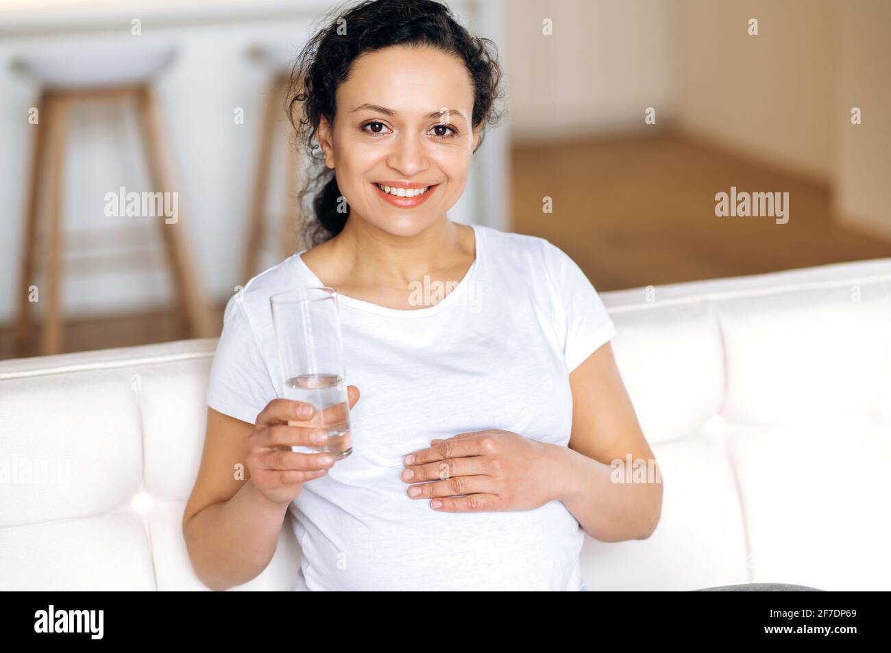 Ziemlich attraktive gesunde Mischling Frau zu Hause im Wohnzimmer sitzen, leben einen gesunden Lebensstil, trinken täglich Menge an Wasser, hält ein Glas sauberes Wasser, Blick auf die Kamera, lächelnd Stockfoto