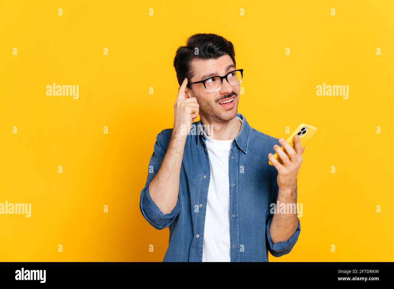 Verwirrt verwirrt verwirrt verwirrt kaukasischen unhaved Kerl, stilvoll gekleidet, hält ein Smartphone in der Hand, Blick pensiv zur Seite, stehen auf einem isolierten orangen Hintergrund Stockfoto