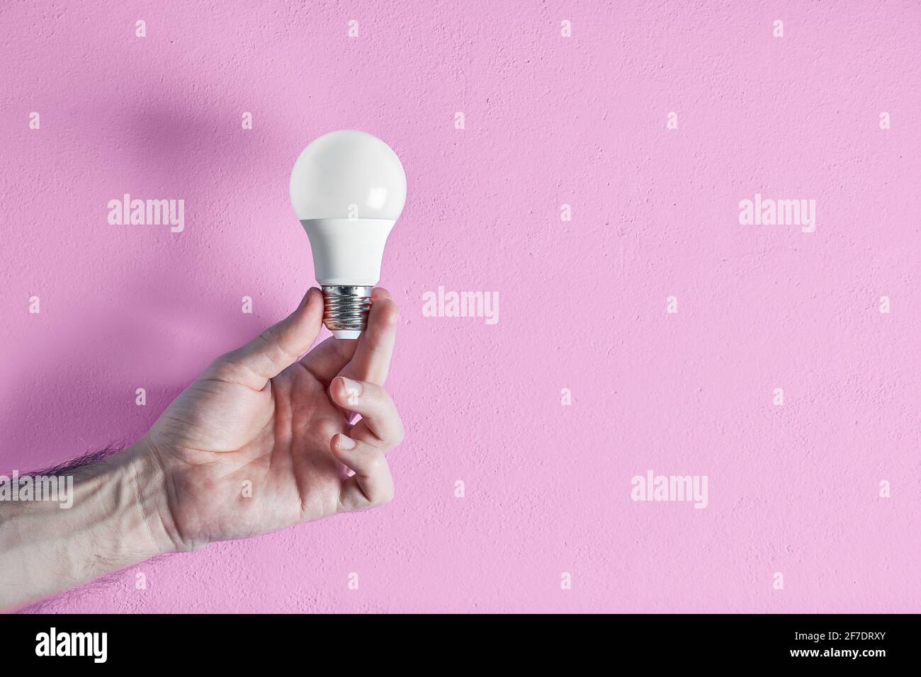 Die Hand eines jungen erwachsenen Mannes hält eine energieeffiziente Glühbirne vor einem rosa Wandhintergrund. Konzept von Kreativität, Effizienz und Nachhaltigkeit. Stockfoto