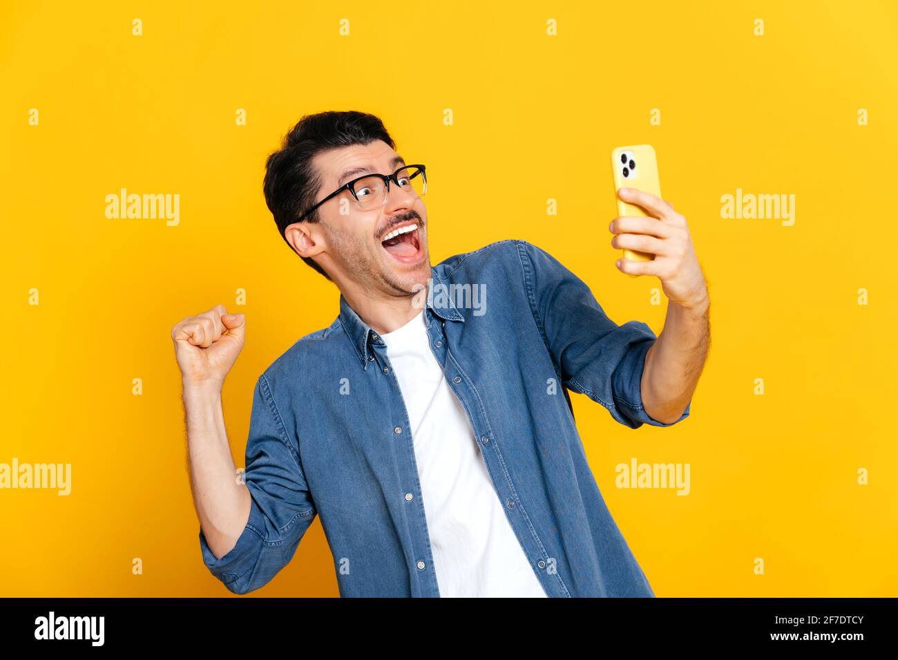 Glücklich fröhlich aufgeregt stilvolle kaukasischen Kerl hält Smartphone, erhalten unerwartete Nachrichten, gewinnen, steht auf isolierten orangefarbenen Hintergrund, fröhlichen Gesichtsausdruck, toothy Lächeln, gestikulieren mit Faust Stockfoto
