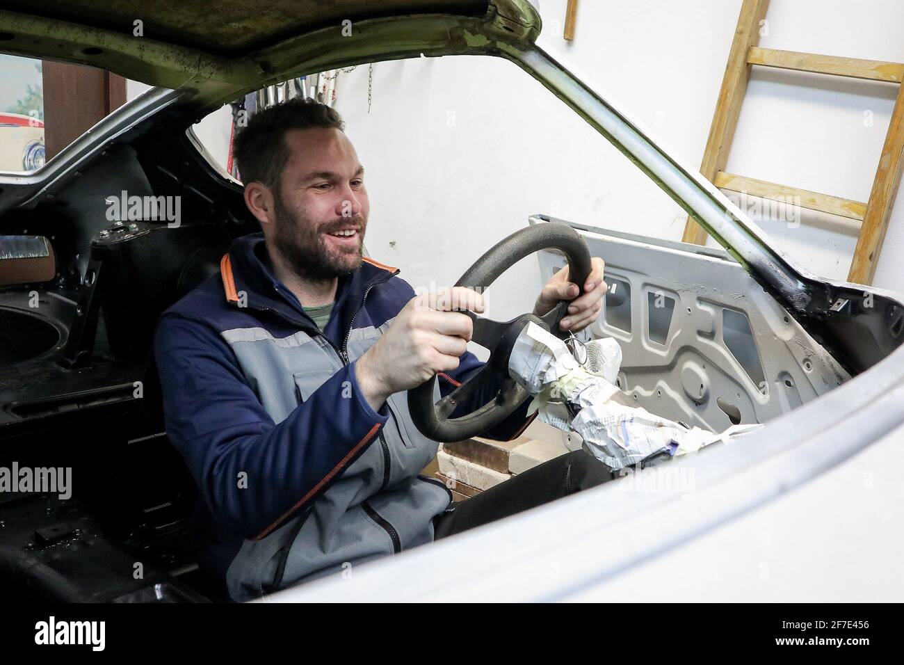 Ein junger Mann sitzt glücklich hinter dem Steuer eines Oldtimers, das gerade restauriert wird. Abgestreiftes Auto bereit zum Malen, ein Mann Traum kommen t Stockfoto