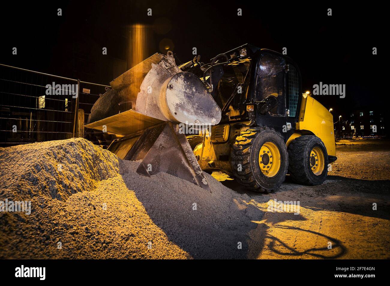 Baustelle an einer Stadtstraße. Ein kleiner, gelber Bagger oder Bobcat, der nachts auf einer Baustelle geparkt hat. Häuser im Hintergrund Stockfoto