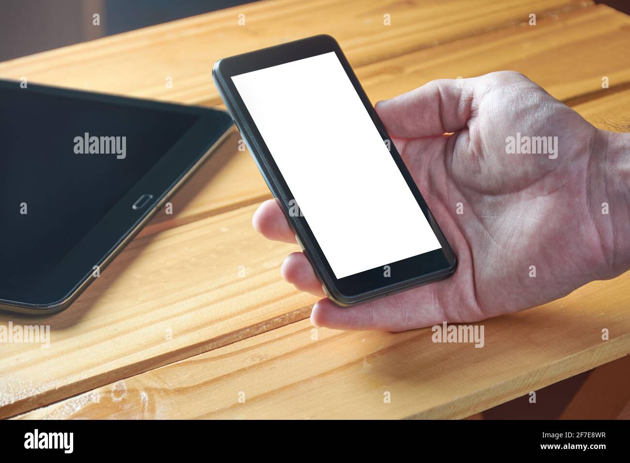 Die Hand eines jungen erwachsenen Mannes hält ein schwarzes Mobiltelefon mit weißem leerem Bildschirm auf einem rustikalen Holztisch. Technologie, Kommunikation, Heimarbeit. Stockfoto