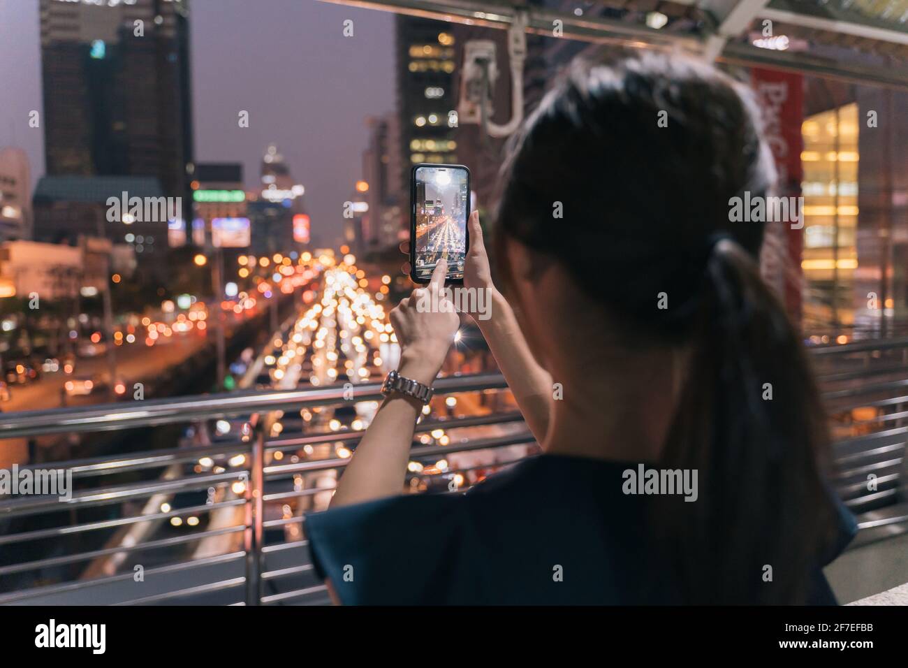 Rückansicht einer jungen, nicht erkennbaren Frau, die auf einer Fußgängerbrücke steht und dabei mitnimmt Bild der Abendansicht der Stadtstraße mit schönem Himmel Anzeigen Stockfoto