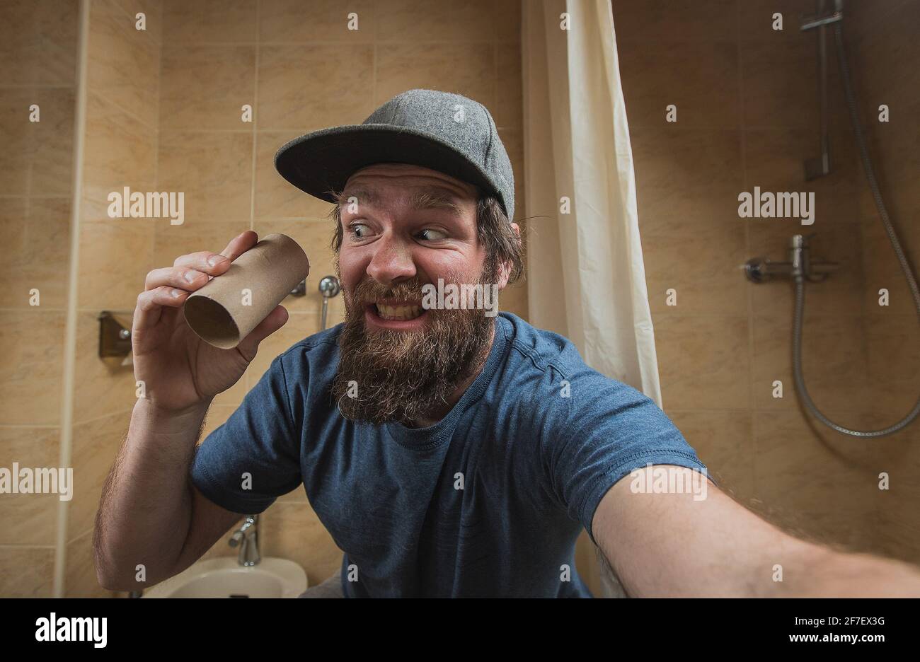 Hipster Mann mit einem verzweifelten oder gestörten Gesicht, als er eine leere Rolle Toilettenpapier im Badezimmer hält. Erschrocken Mann von laufen aus Toilettenpapier. Stockfoto