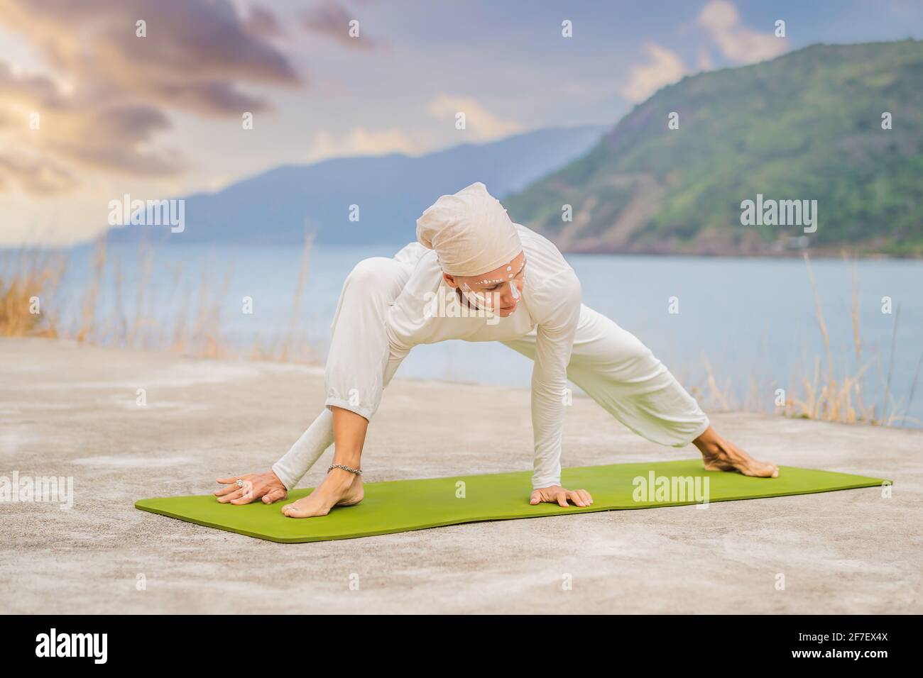 Kundalini Yoga Frau in weißen Kleidern und Turban praktiziert Yoga Kundalini auf dem Hintergrund des Meeres, der Berge und des Sonnenuntergangs. Fighting Face Painting Stockfoto