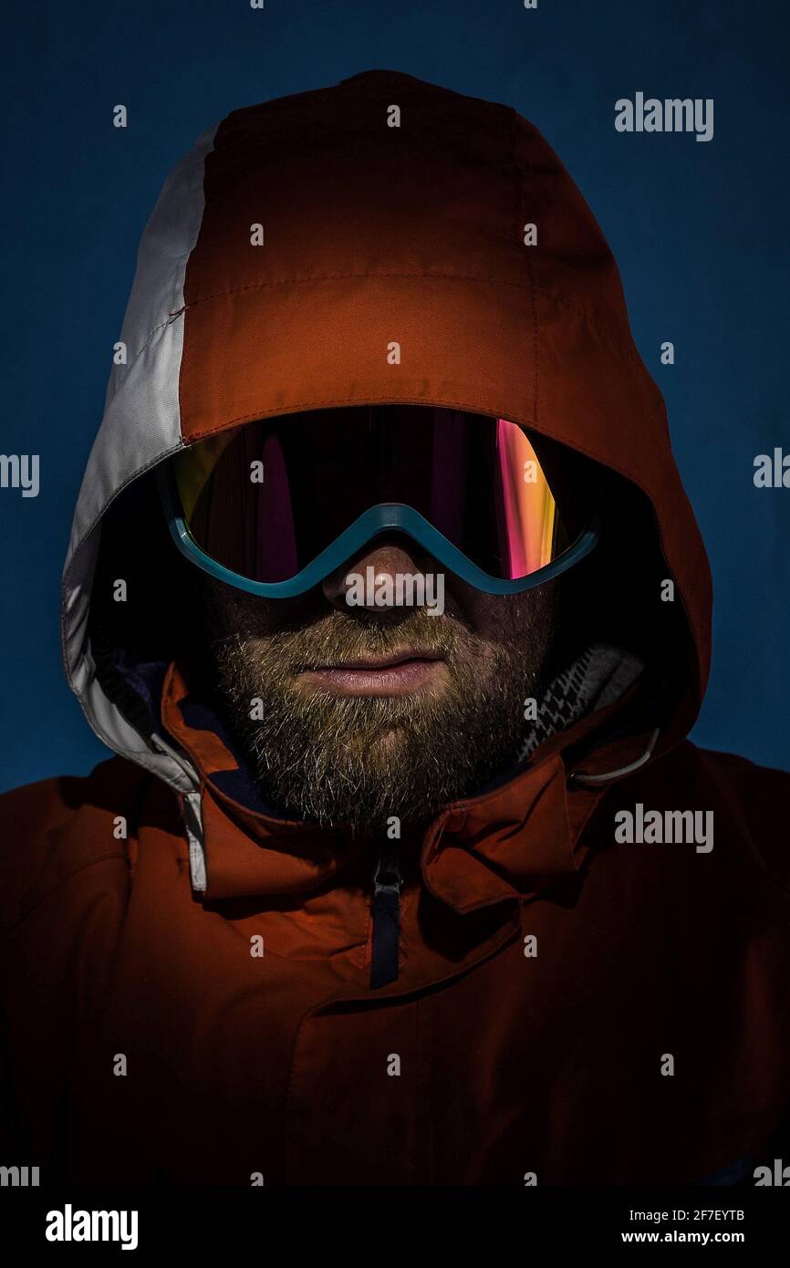 Porträt eines jungen Mannes mit Bart, Grinsen im Gesicht, Skijacke über dem Kopf und Brille auf dunklem Hintergrund. Schnee fällt. Konzept eines Snowboards Stockfoto