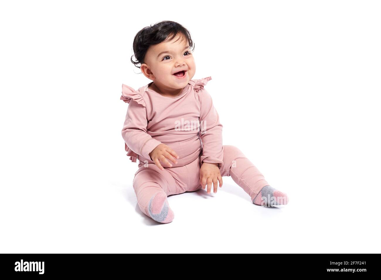 Entzückende weibliche Baby in schönen rosa Outfit lächelnd mit Mund offen. Attraktives kleines Kind, das auf dem Boden sitzt und posiert, isoliert auf weißem Studiohintergrund. Konzept der Kindheit. Stockfoto
